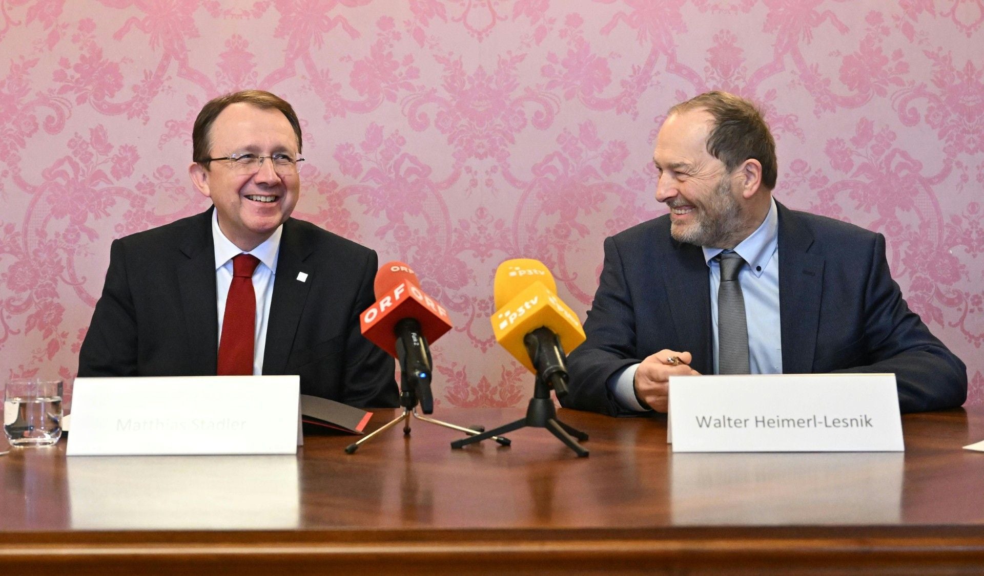 Zwei Männer im Anzug sitzen bei einer Pressekonferenz mit Mikrofonen und Namensschildern.