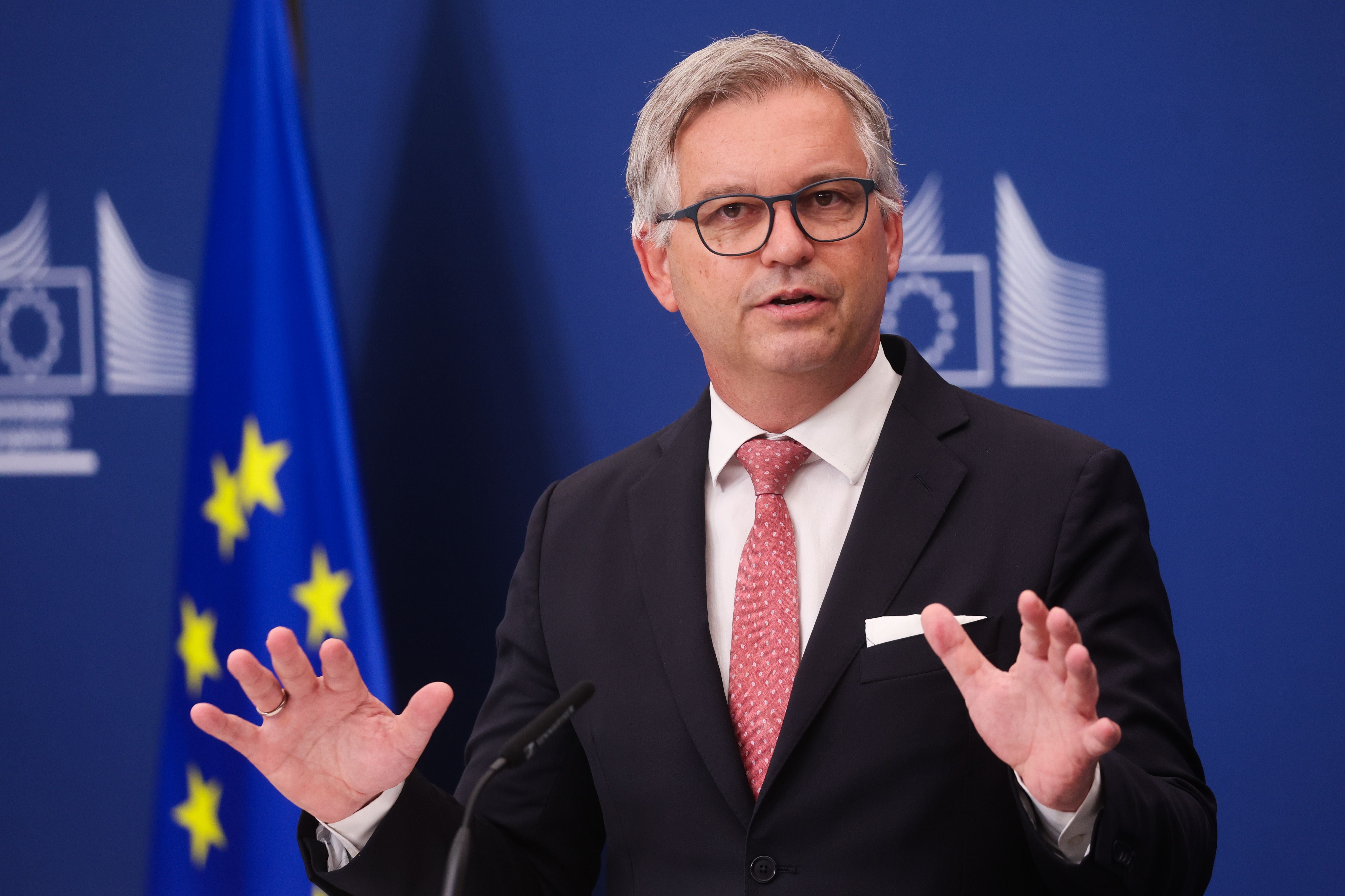 Ein Mann im Anzug spricht bei einer Pressekonferenz vor einer EU-Flagge und blauem Hintergrund.