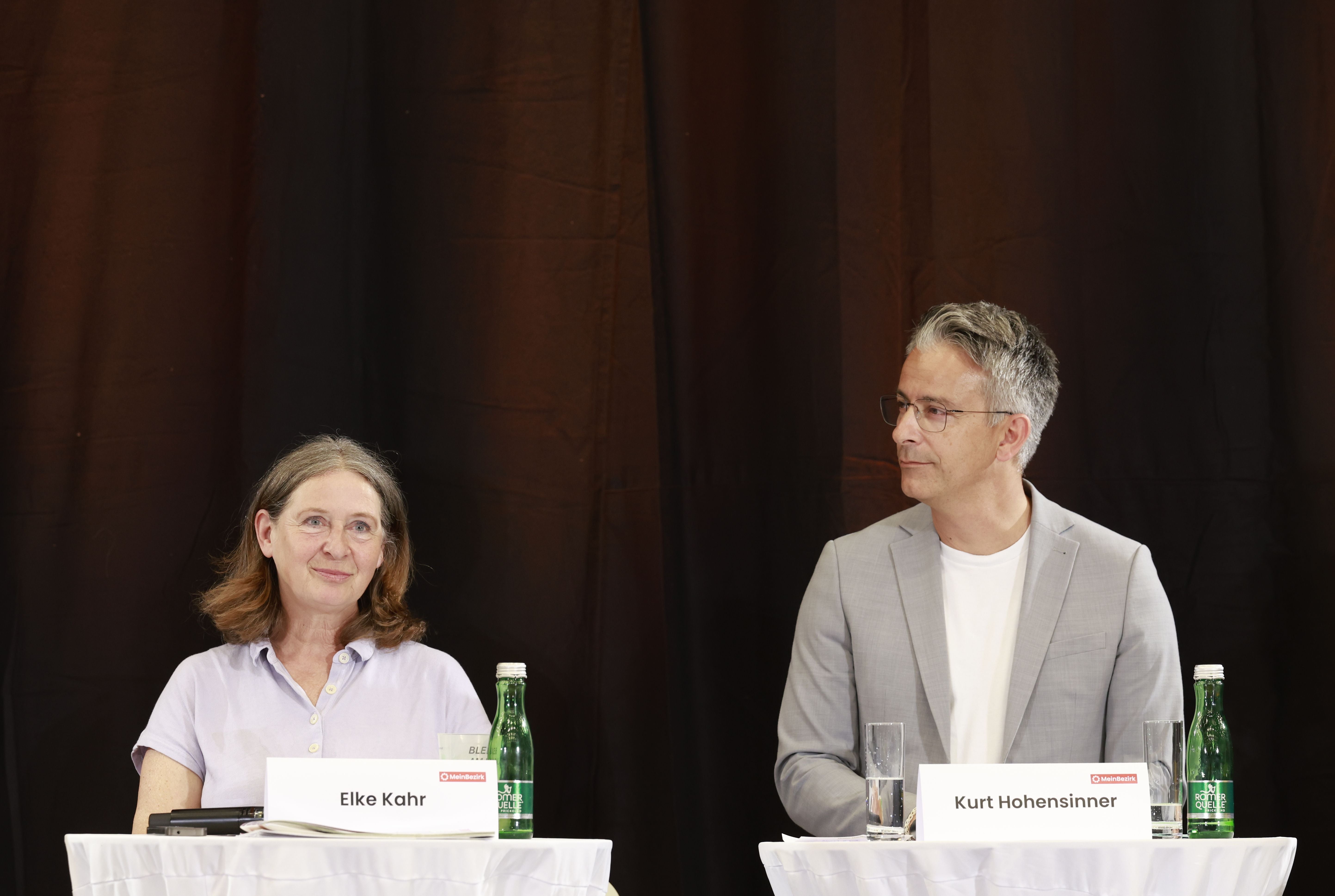 Zwei Personen sitzen an Tischen mit Namensschildern vor dunklem Hintergrund bei einer Podiumsdiskussion.