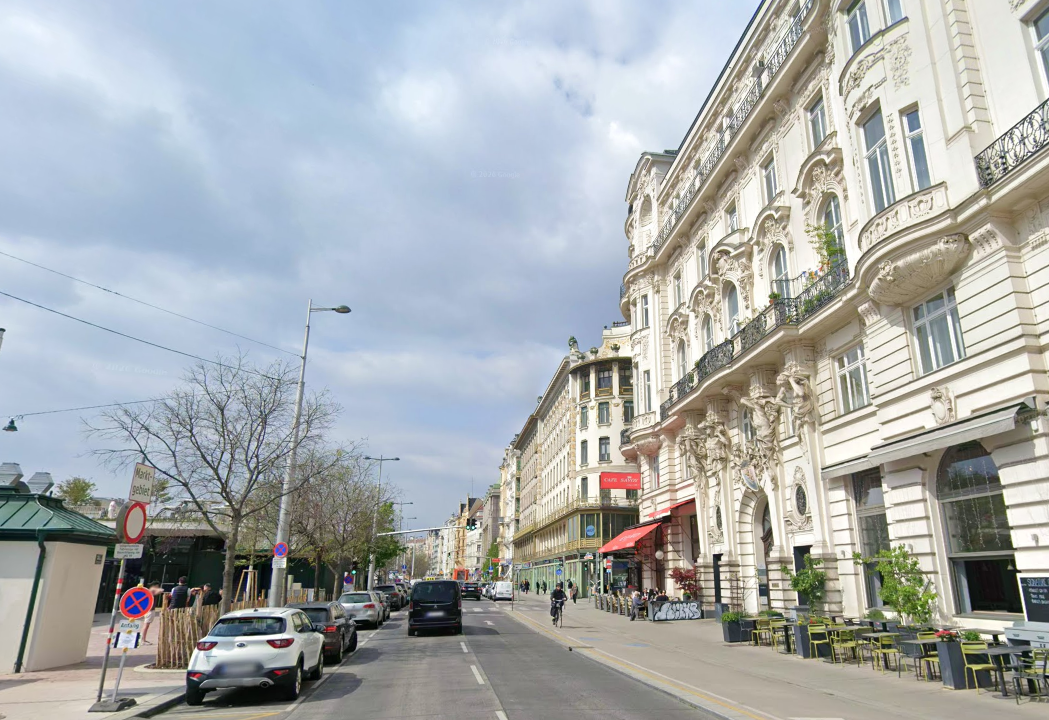 Straßenszene mit prunkvollen Gründerzeithäusern, Cafés und geparkten Autos bei bewölktem Himmel.