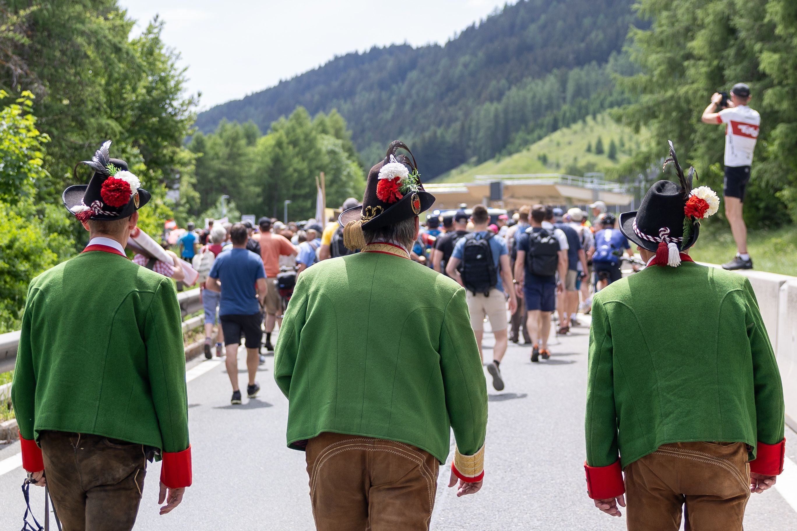 Drei Männer in Tiroler Tracht gehen bei einer Demo auf der Brennerautobahn Richtung Menschenmenge.