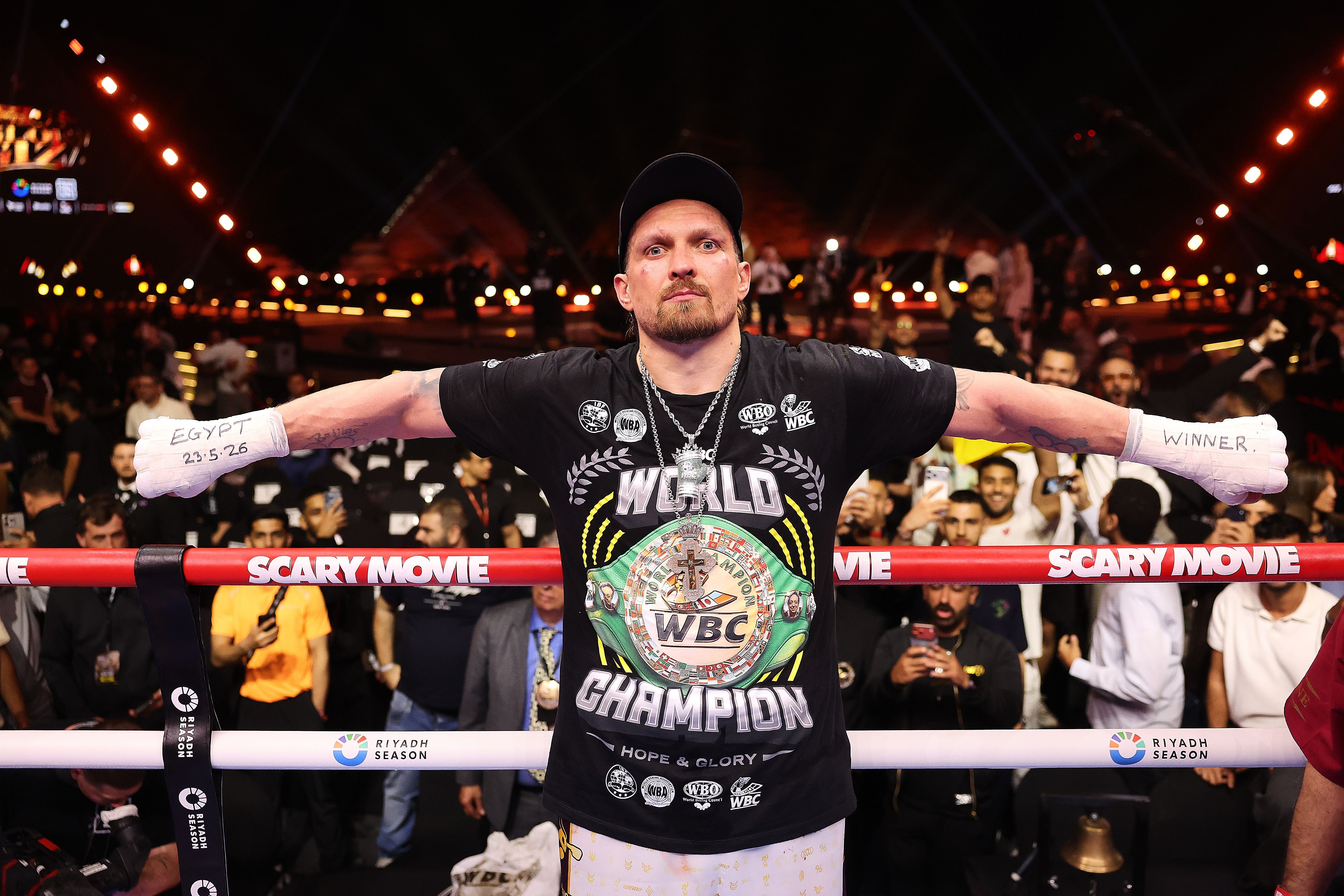 Ein Boxer mit einem "World Champion" WBC-Shirt streckt die Arme im Boxring aus, jubelnde Zuschauer im Hintergrund.