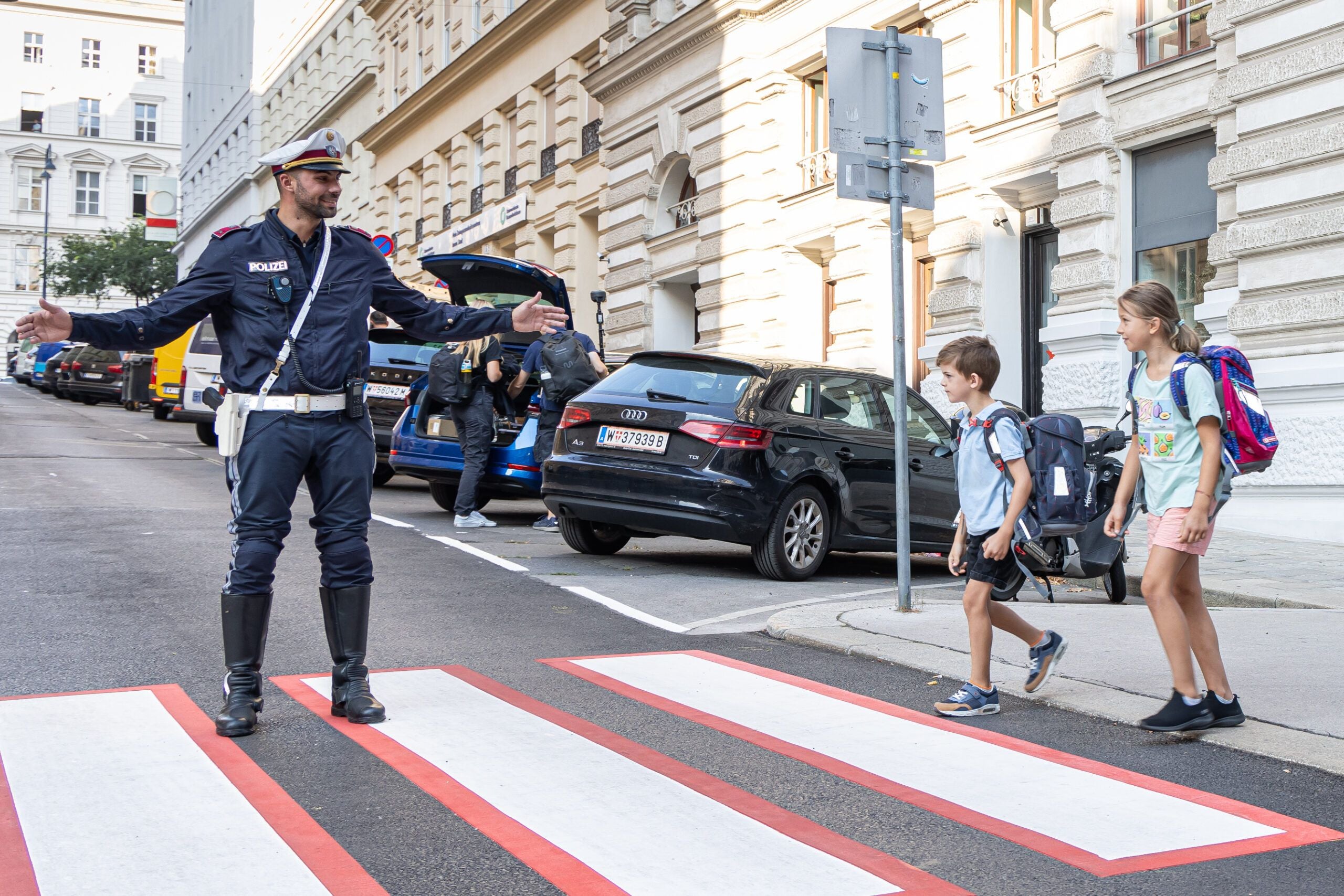 Ein Polizist hilft zwei Schulkindern beim Überqueren eines Zebrastreifens vor einer Schule.