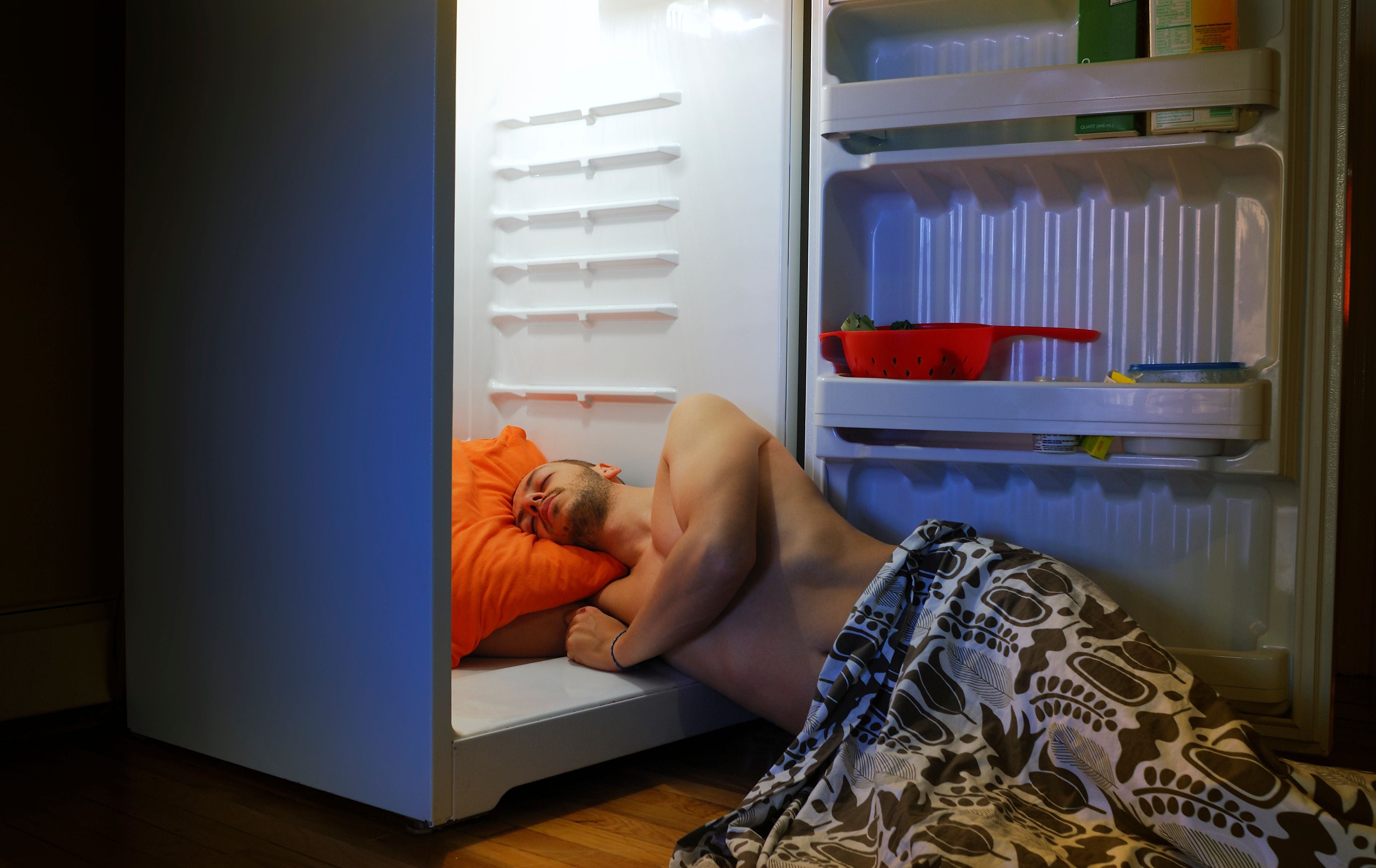 Ein Mann schläft mit Kopfkissen und Decke im offenen, leeren Kühlschrank.