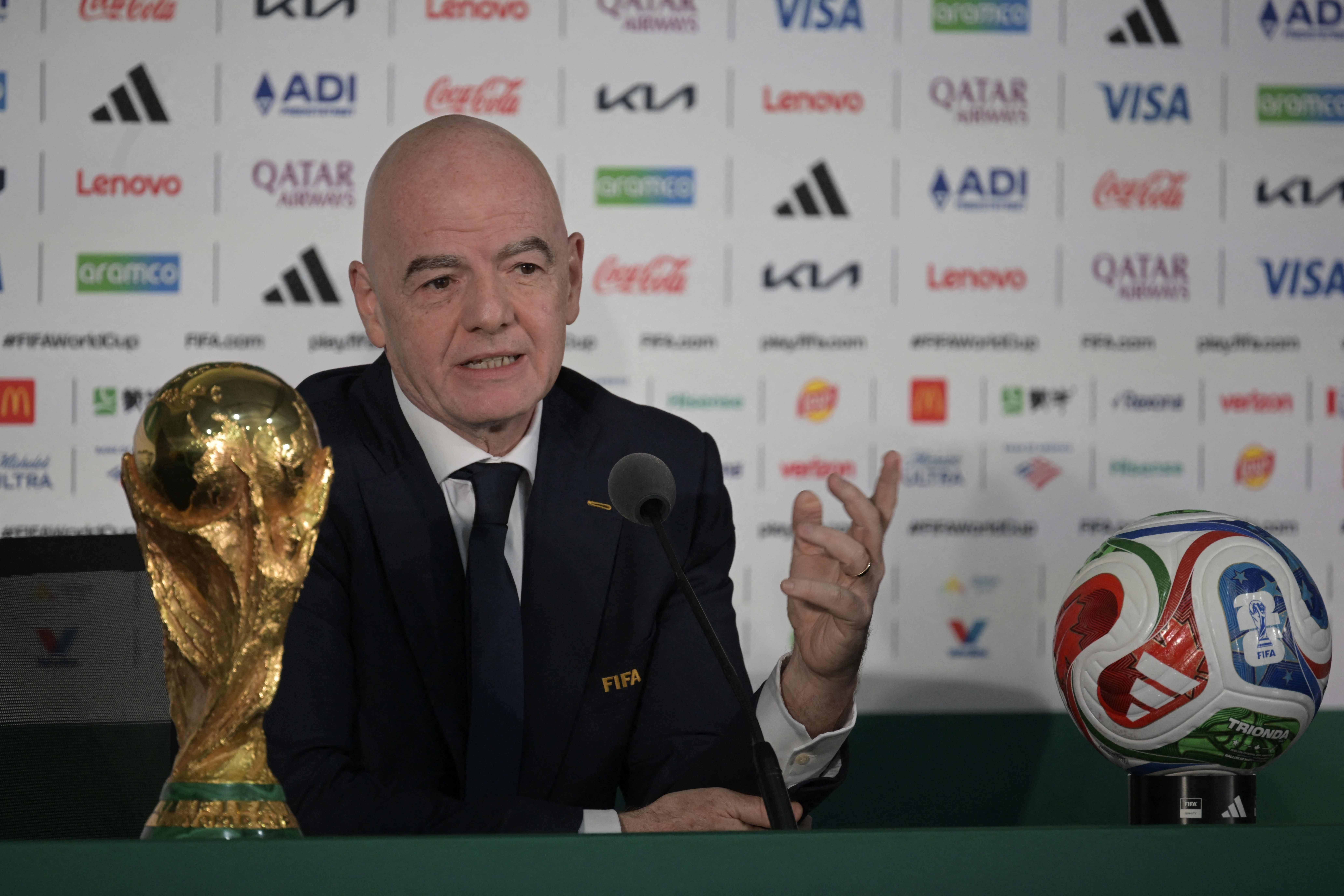 Ein Mann im Anzug sitzt vor Mikrofon, WM-Pokal und Fußball bei einer Pressekonferenz.