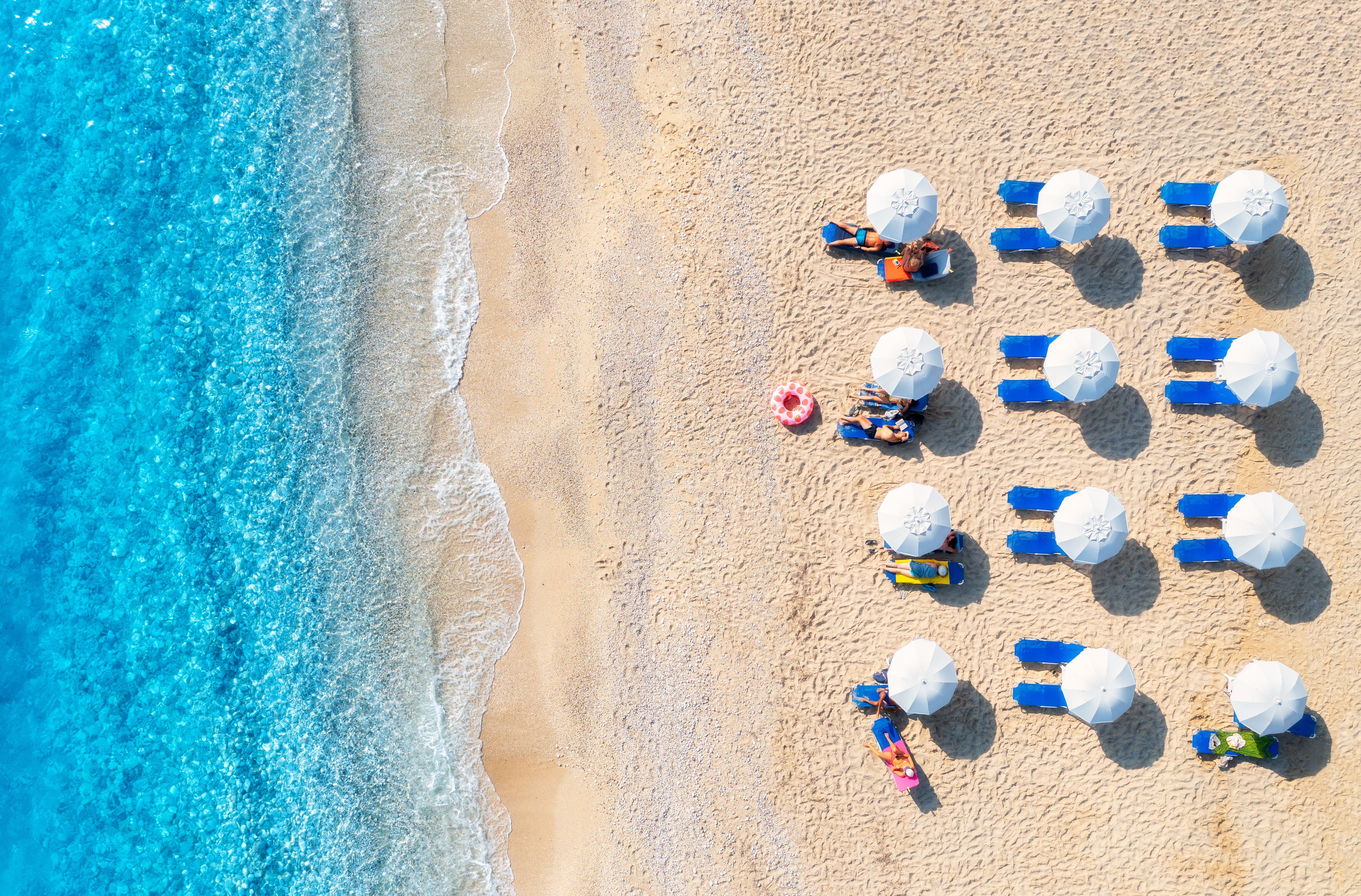 Luftaufnahme eines Strandes mit Sonnenschirmen, Liegen und türkisblauem Meer in Griechenland.
