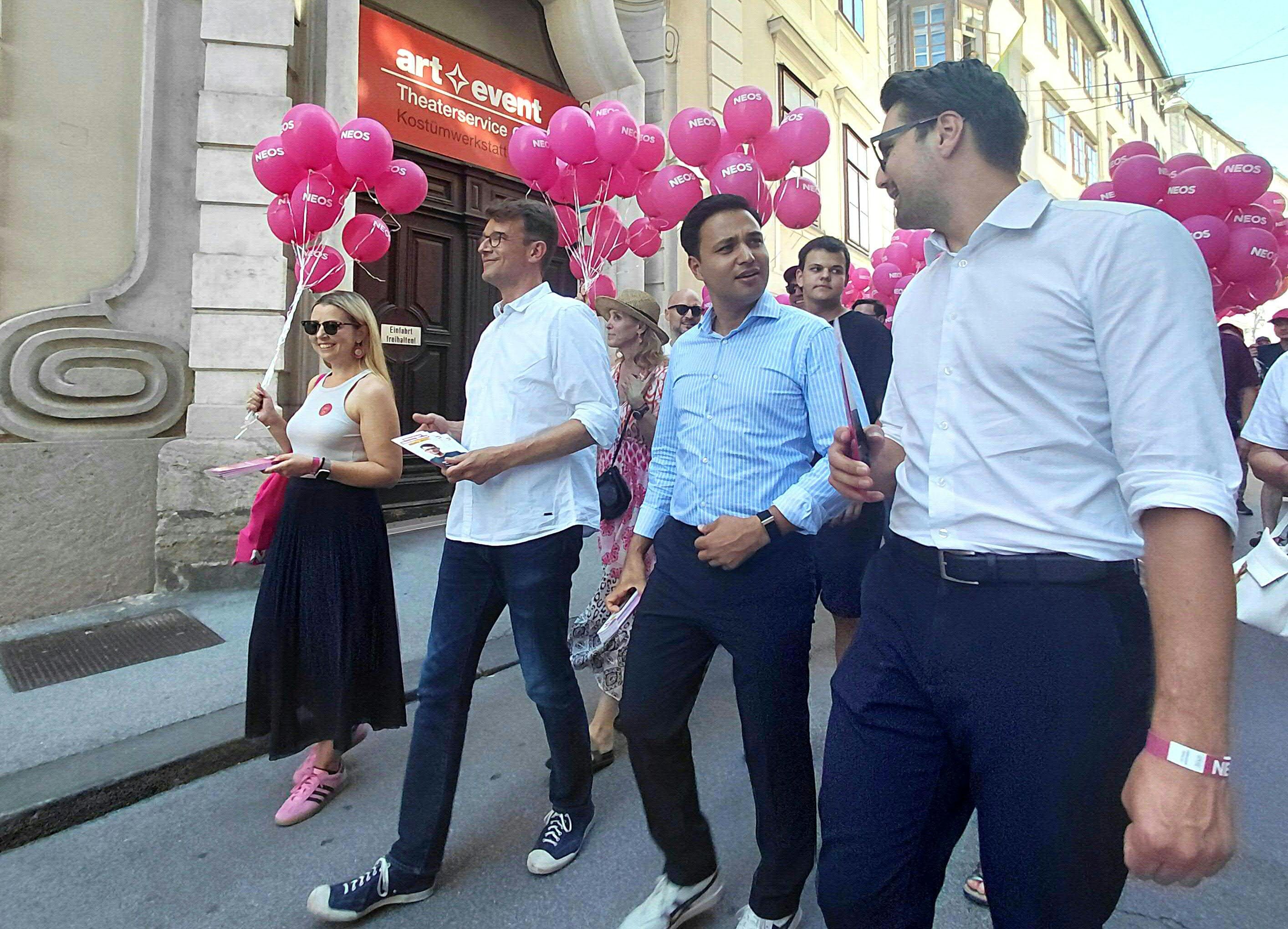 Mehrere Menschen mit NEOS-Luftballons gehen bei einer Wahlkampfveranstaltung in einer Stadtstraße.