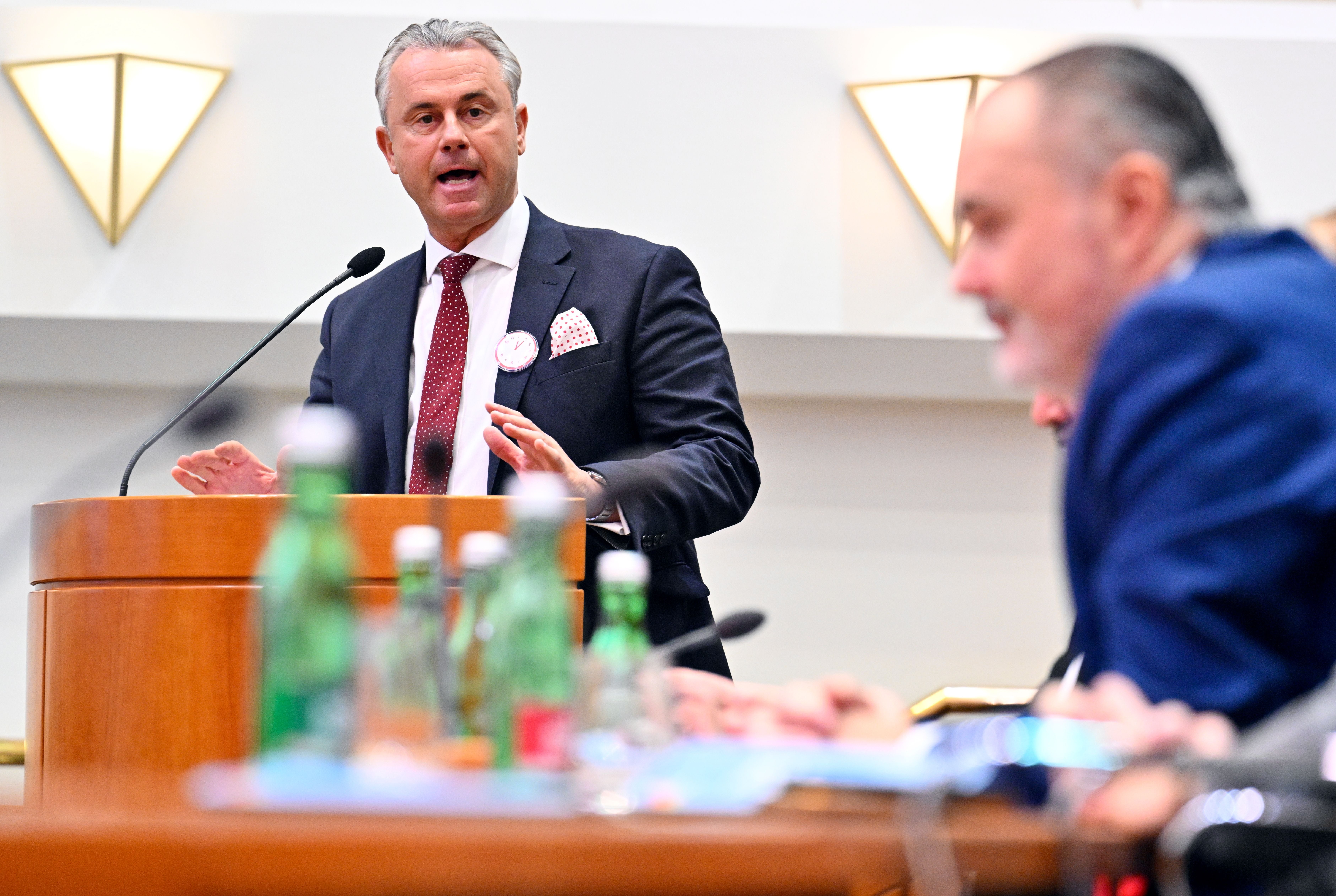 Ein Mann im Anzug spricht an einem Rednerpult bei einer Sitzung, weitere Personen im Vordergrund.
