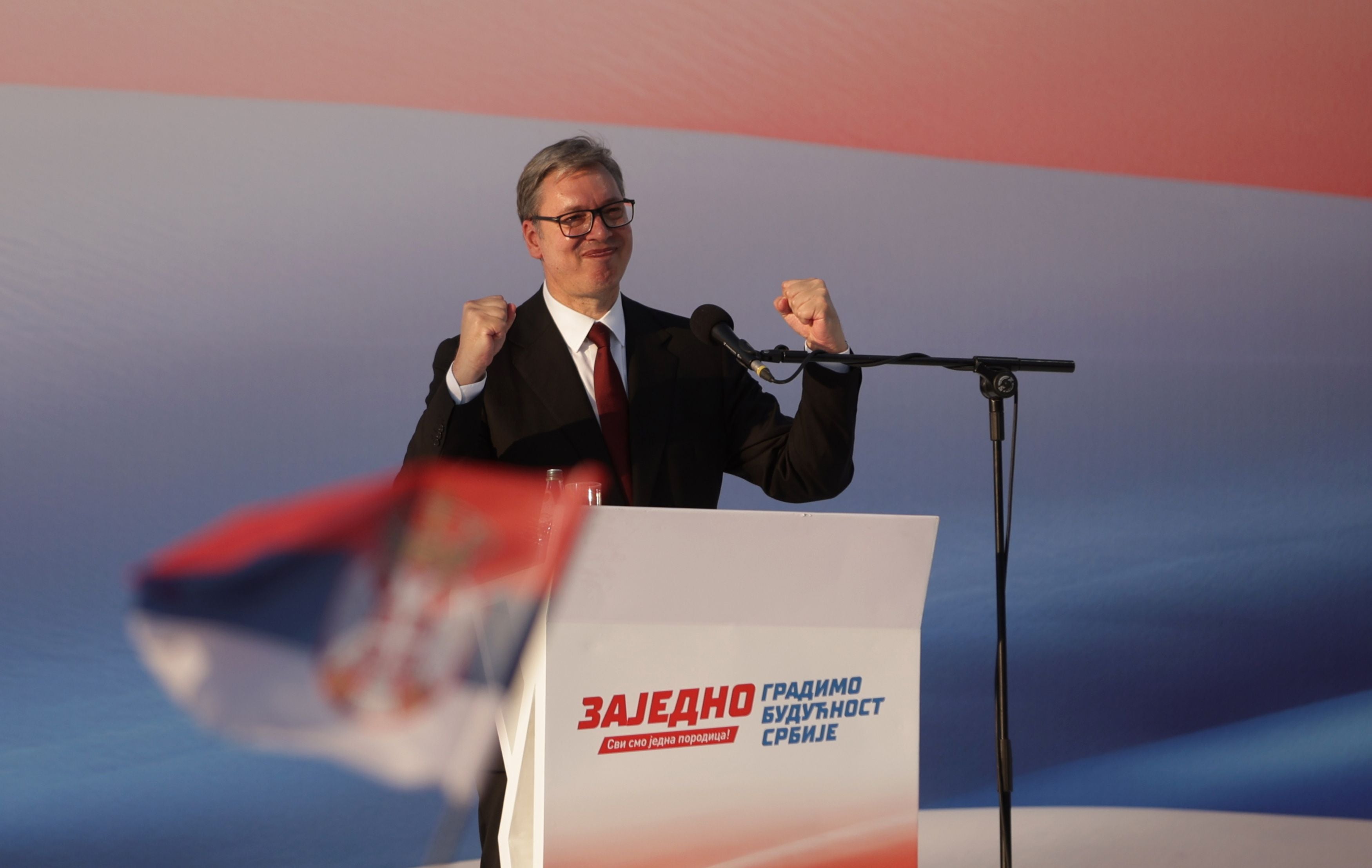 Ein Mann im Anzug gestikuliert an einem Rednerpult mit serbischer Flagge im Vordergrund.