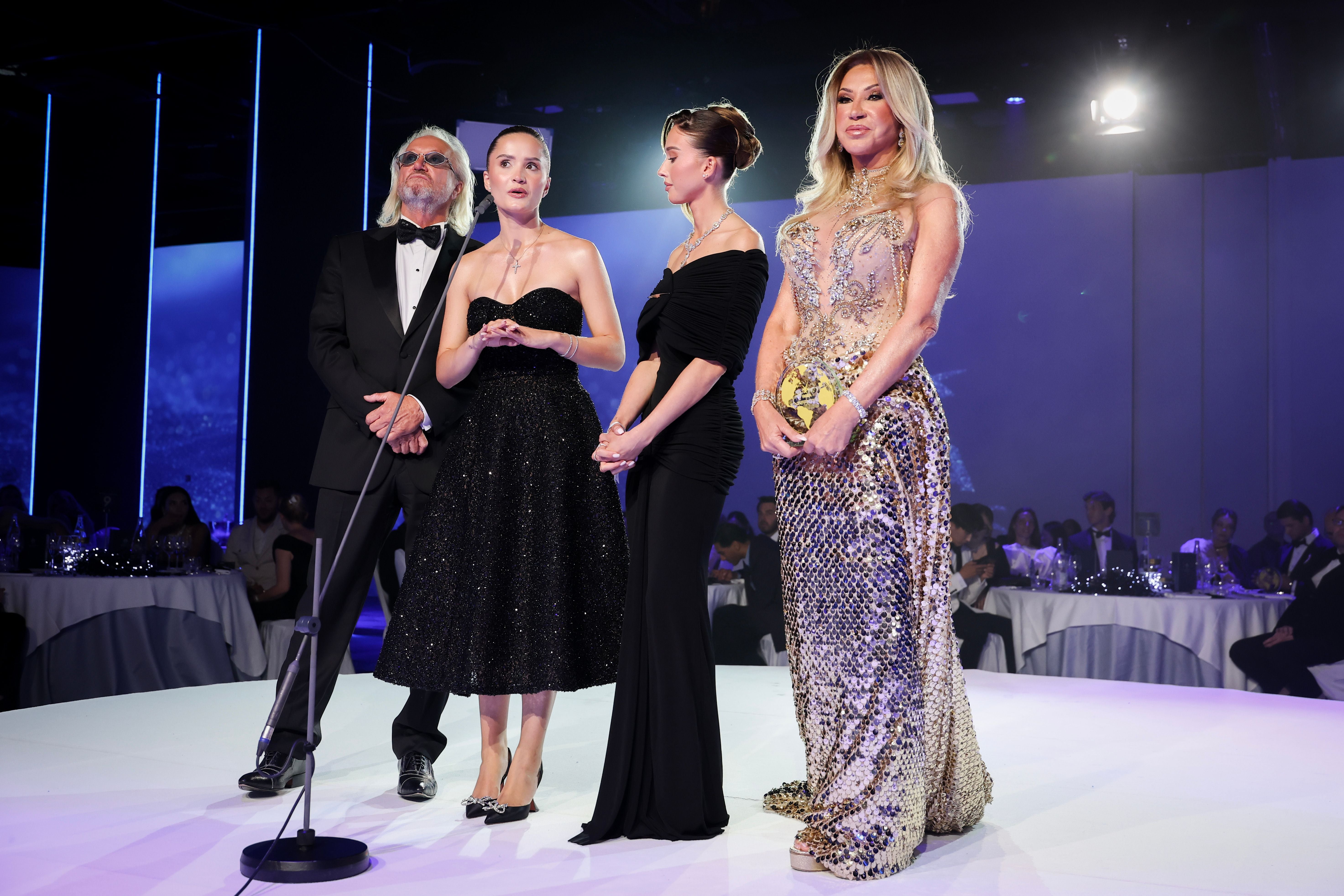 Vier elegant gekleidete Personen stehen auf einer Bühne bei einer Gala mit Publikum im Hintergrund.
