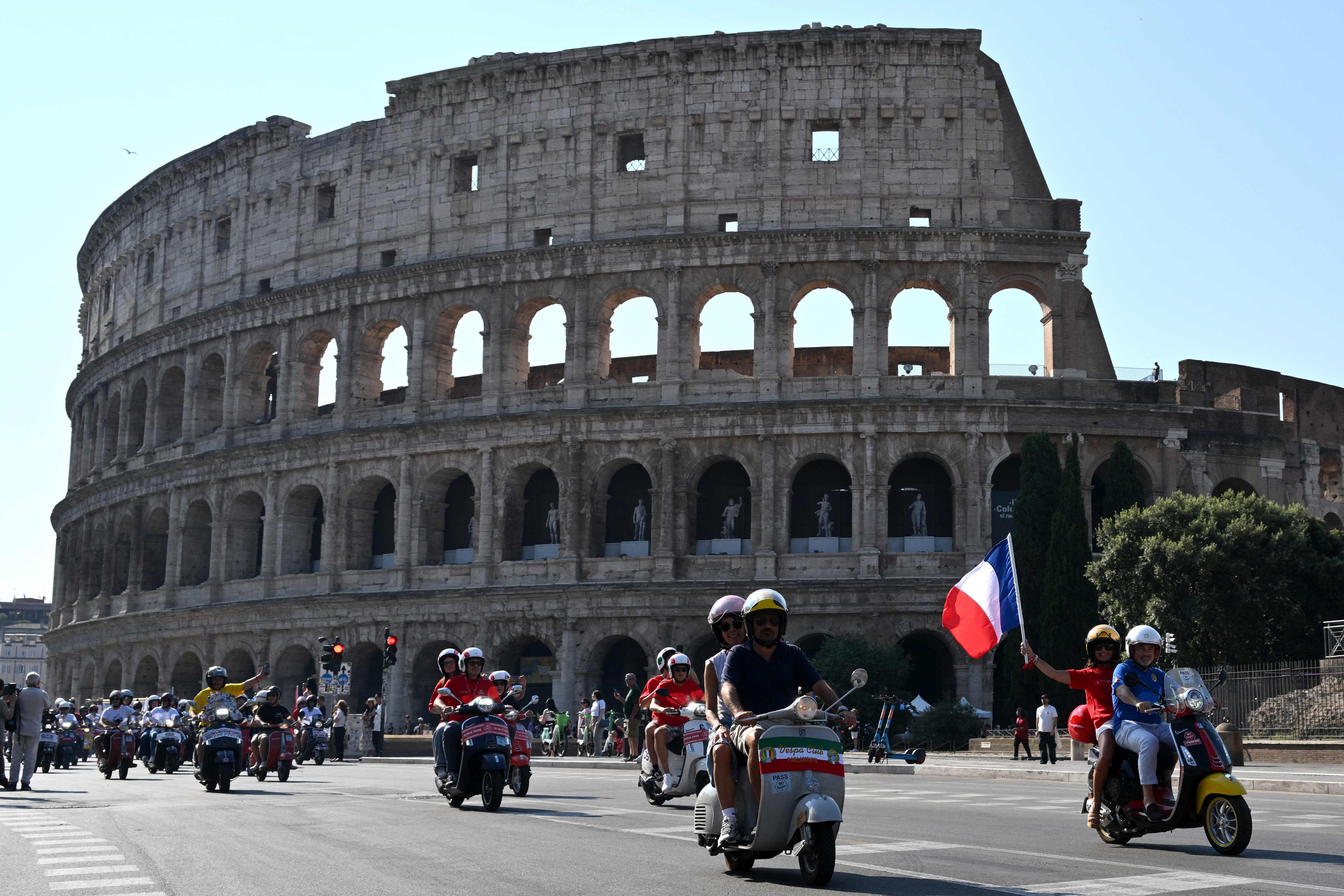 Mehrere Menschen fahren auf Vespas am Kolosseum in Rom vorbei, eine Person hält eine französische Flagge.