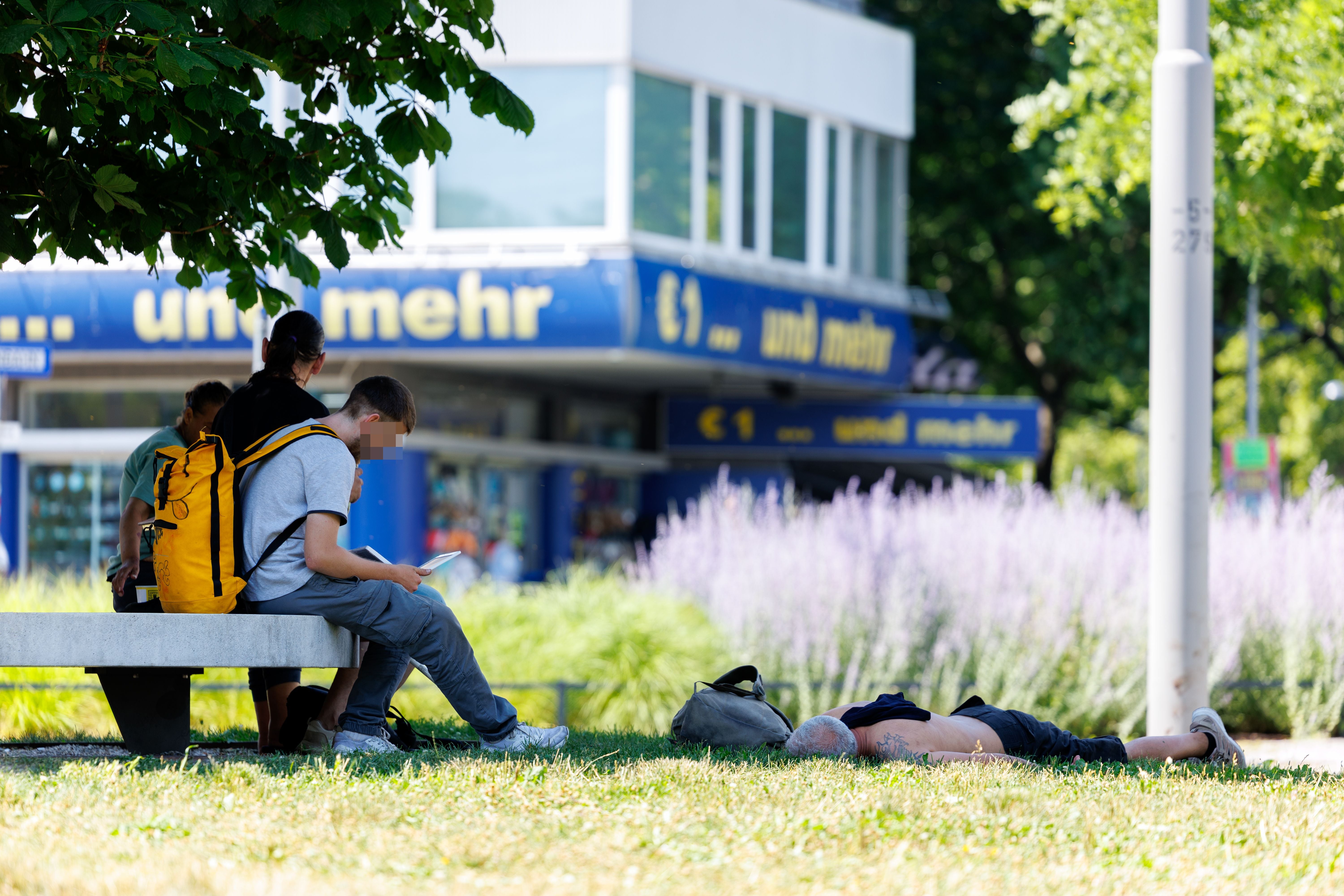 Menschen im Schatten und ein Mann liegend auf Gras während einer sommerlichen Hitzewelle in Wien.