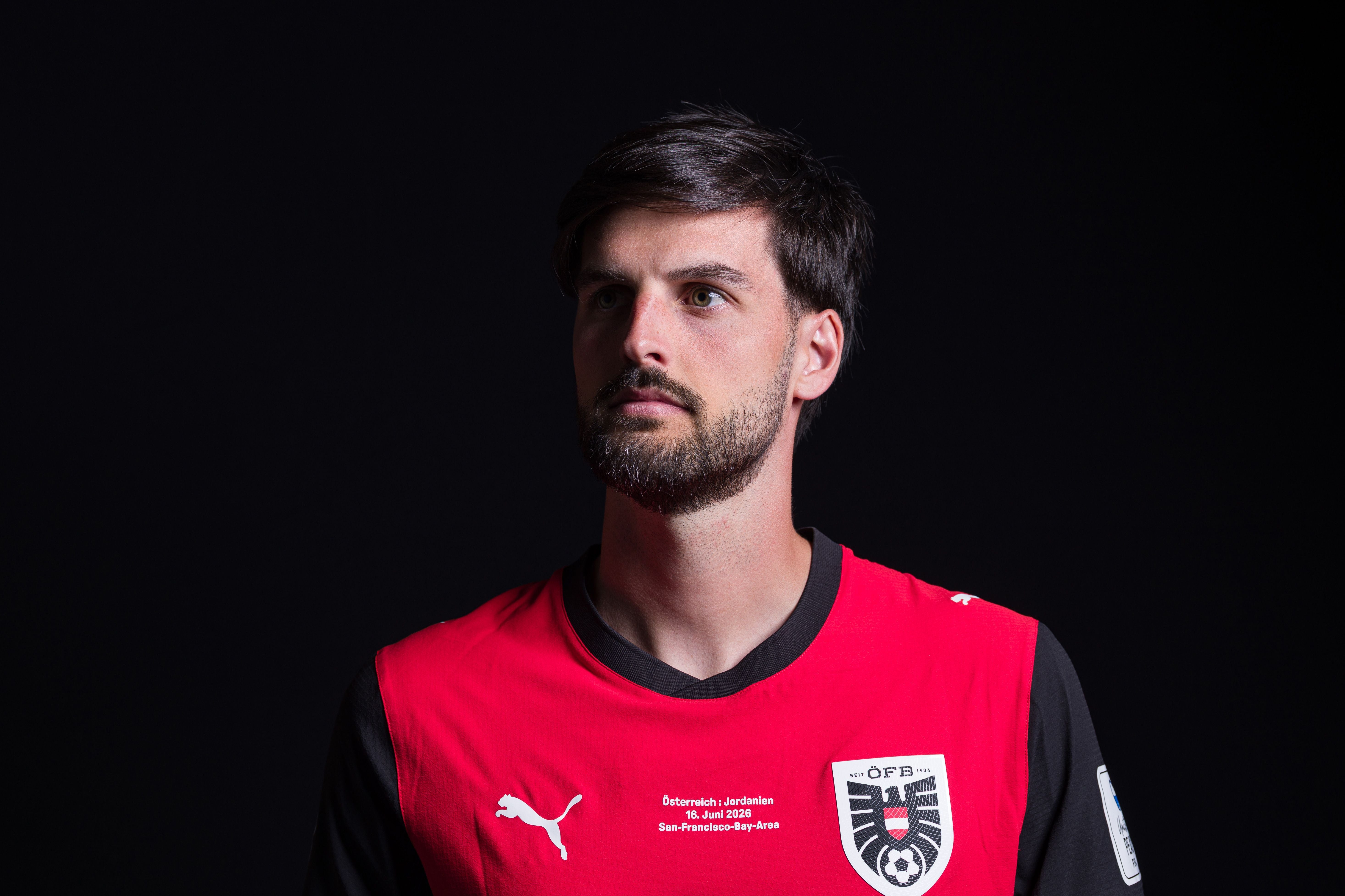 Ein Fußballspieler im roten Trikot posiert vor schwarzem Hintergrund für ein Porträtfoto.