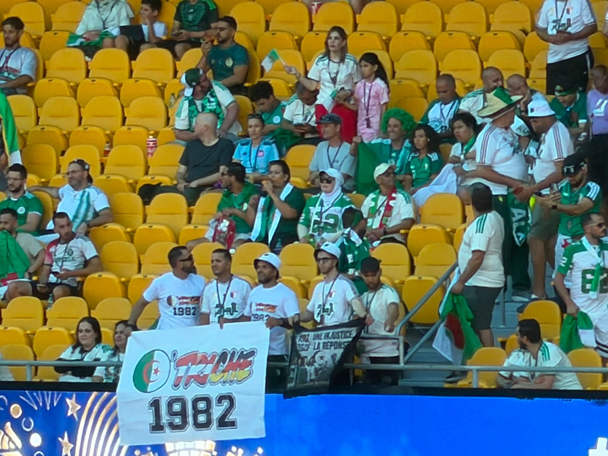 Algerische Fußballfans sitzen in gelben Stadionreihen und halten Fahnen und Banner hoch.