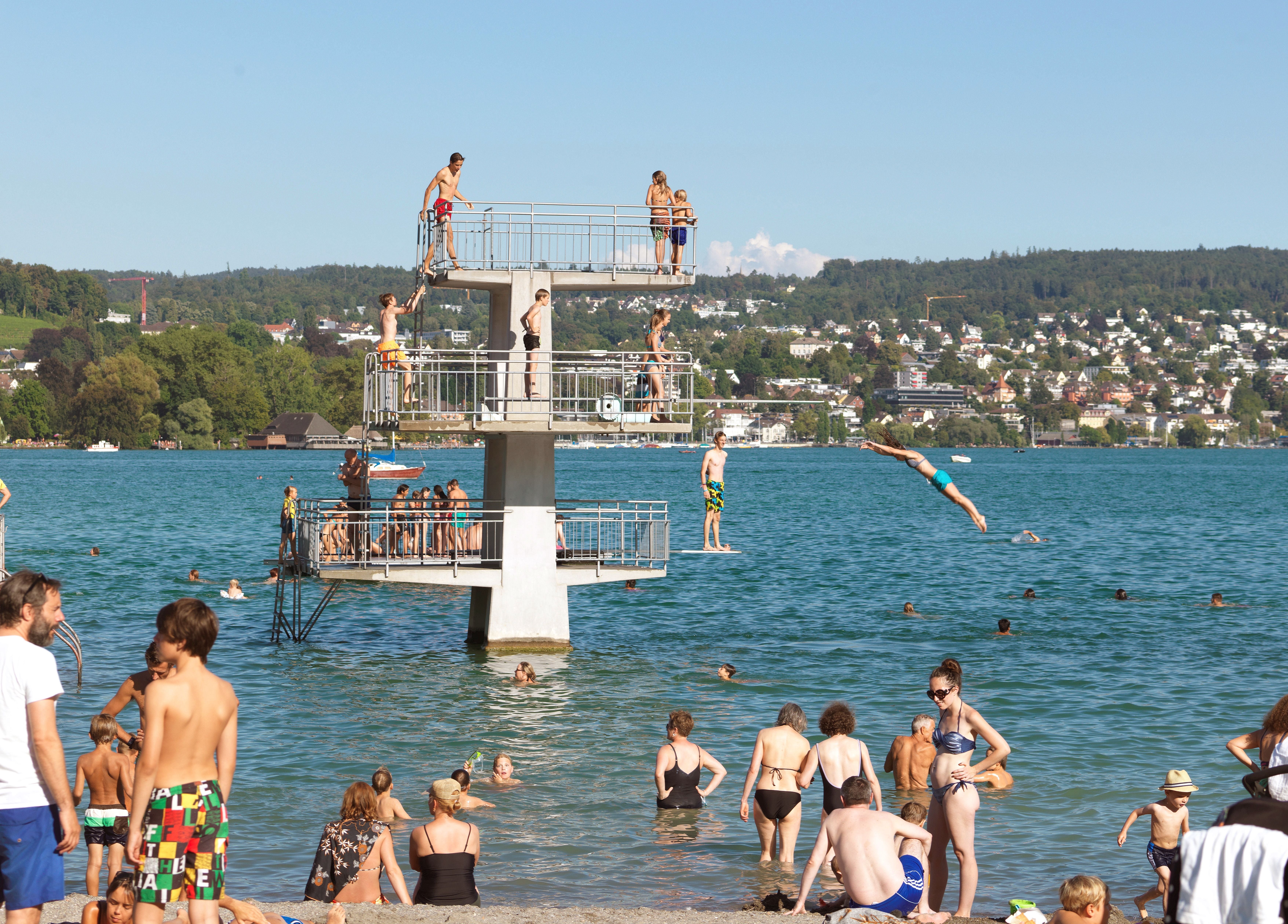 Viele Menschen baden und springen von einem Sprungturm in einen See an einem sonnigen Tag.