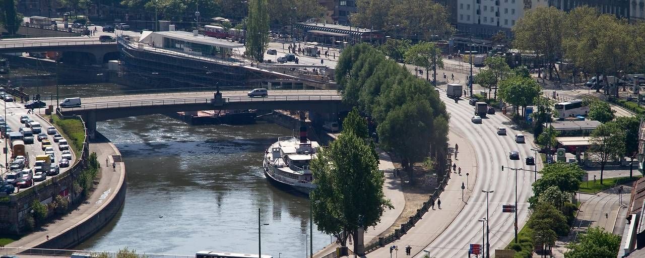 Straße mit Autos und Brücken verläuft entlang eines Flusses, Bäume und ein Schiff sind sichtbar.