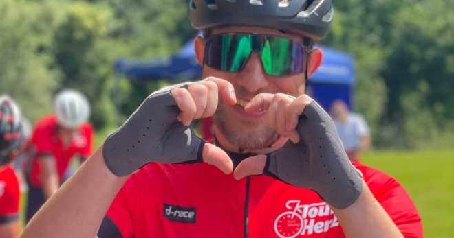Ein Radfahrer formt ein Herz mit den Händen und trägt ein rotes Trikot und Helm.
