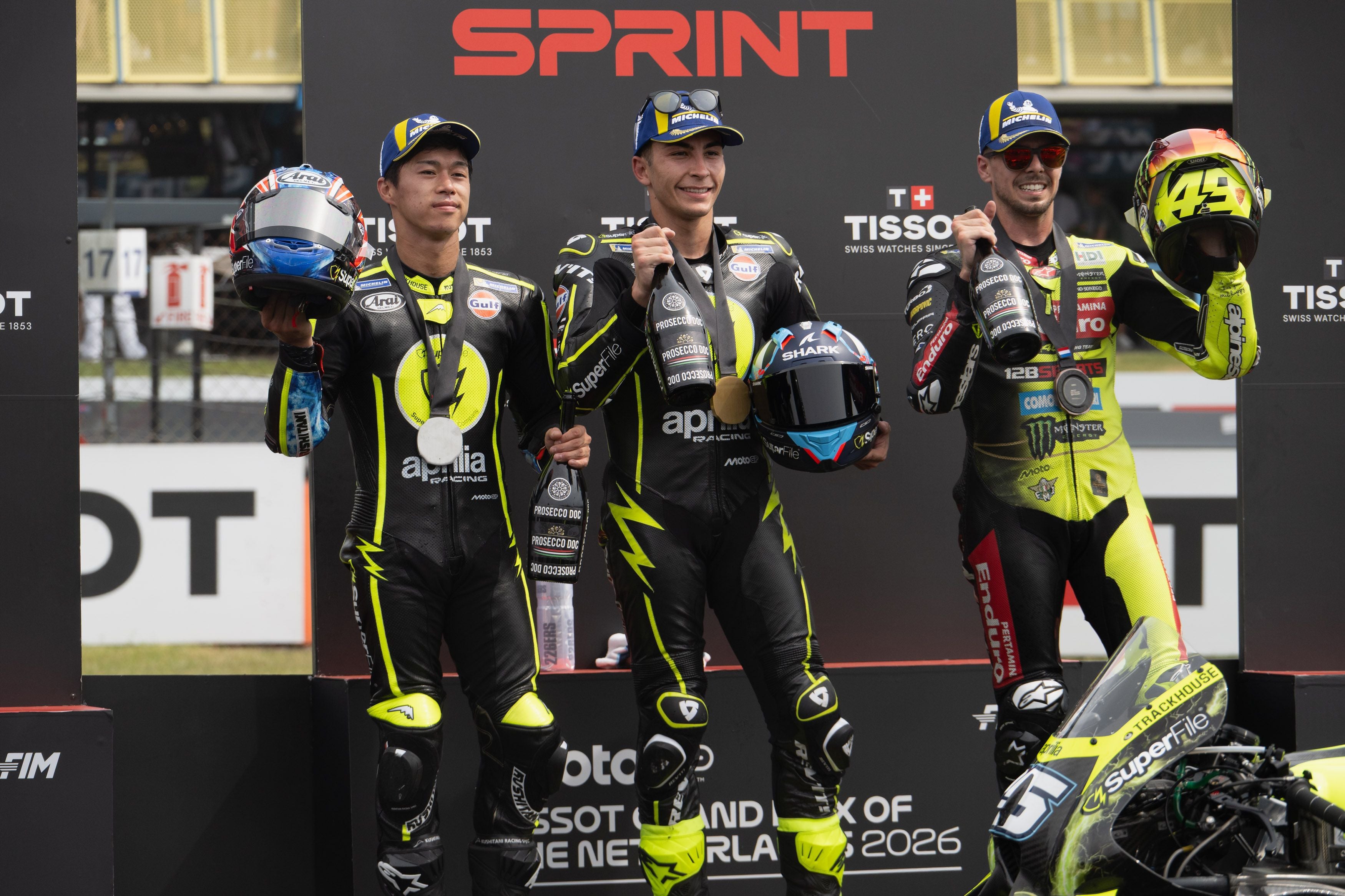 Drei Motorradrennfahrer stehen lächelnd nebeneinander auf einem Podest vor einer schwarzen Wand mit der Aufschrift Sprint. Sie tragen Lederkombis, Kappen, Medaillen um den Hals und halten Champagnerflaschen sowie Helme in den Händen.