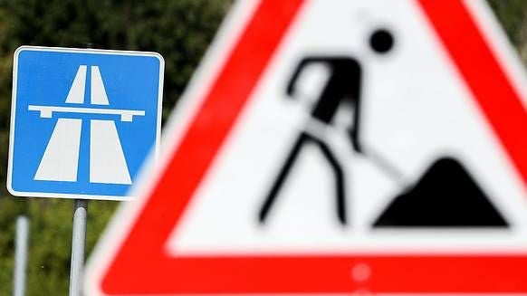 Autobahnschild und unscharfes Baustellenschild im Vordergrund weisen auf Bauarbeiten hin.