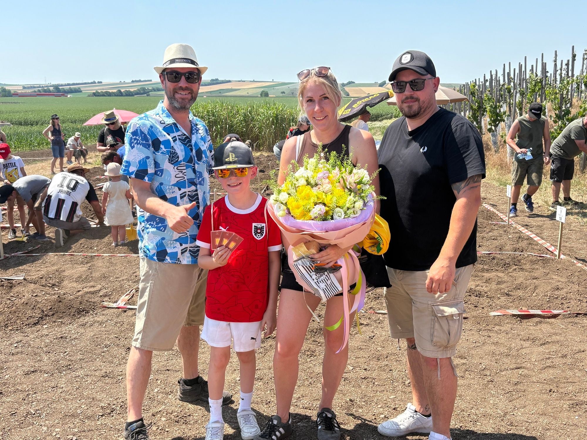Vier Personen, darunter ein Kind, posieren fröhlich im Freien mit einem Blumenstrauß.