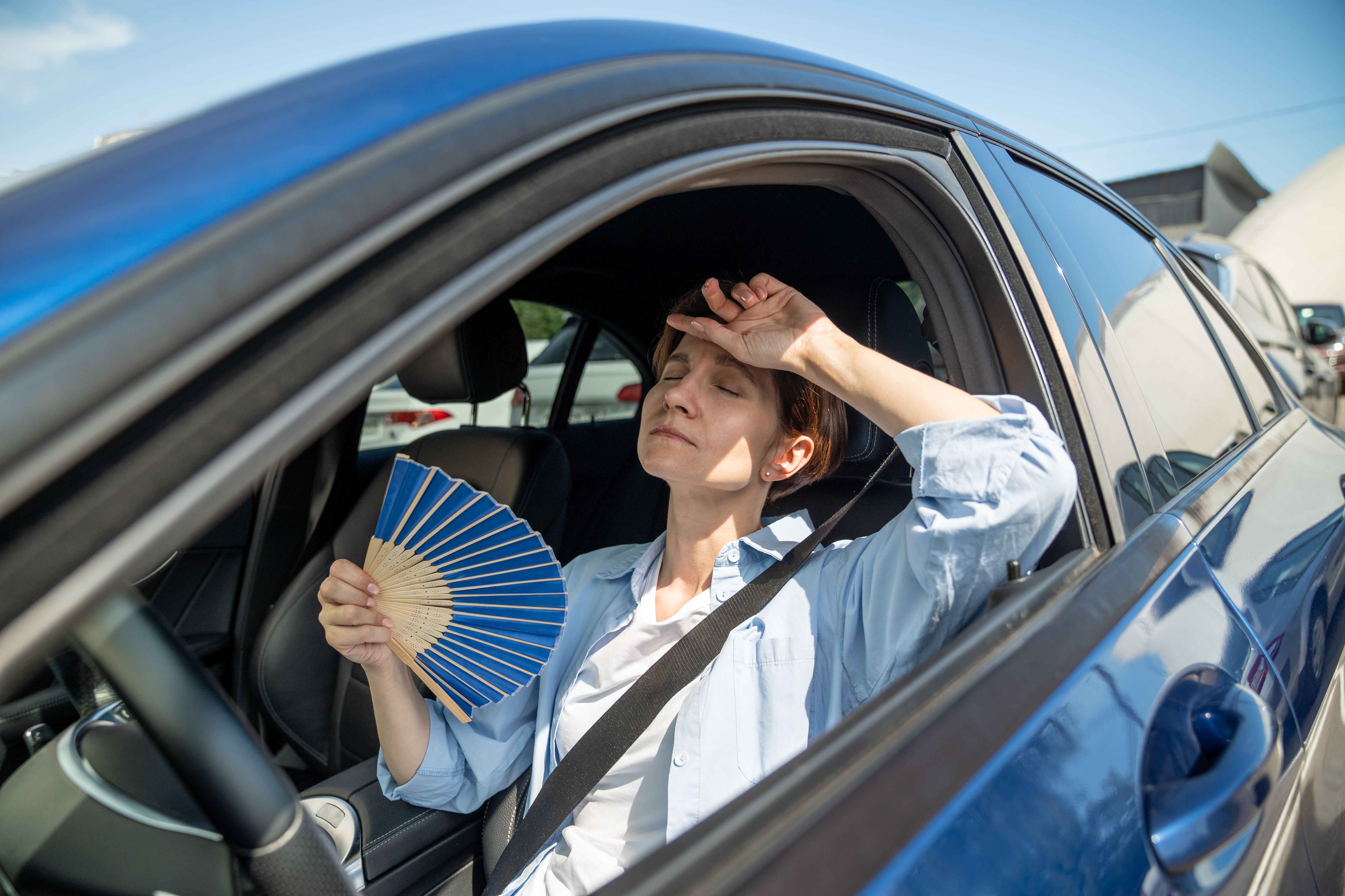 Frau sitzt erschöpft im Auto, fächert sich Luft zu und leidet unter Sommerhitze im Stau.
