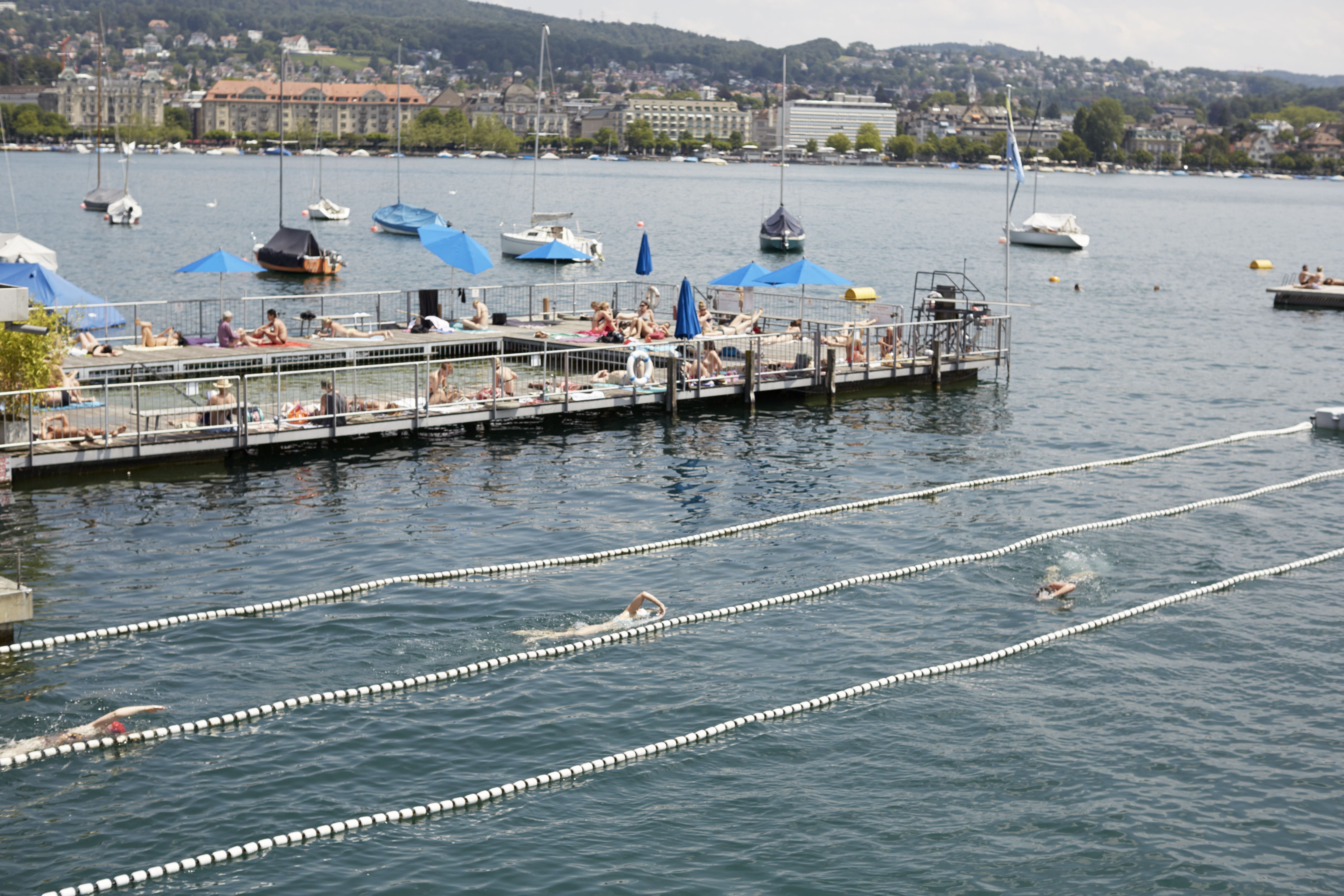 Schwimmer ziehen Bahnen im See, während Menschen auf einem Steg in der Sonne liegen.