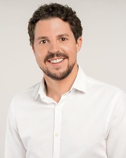 Ein Mann mit lockigem Haar und Bart lächelt, trägt ein weißes Hemd vor hellem Hintergrund.