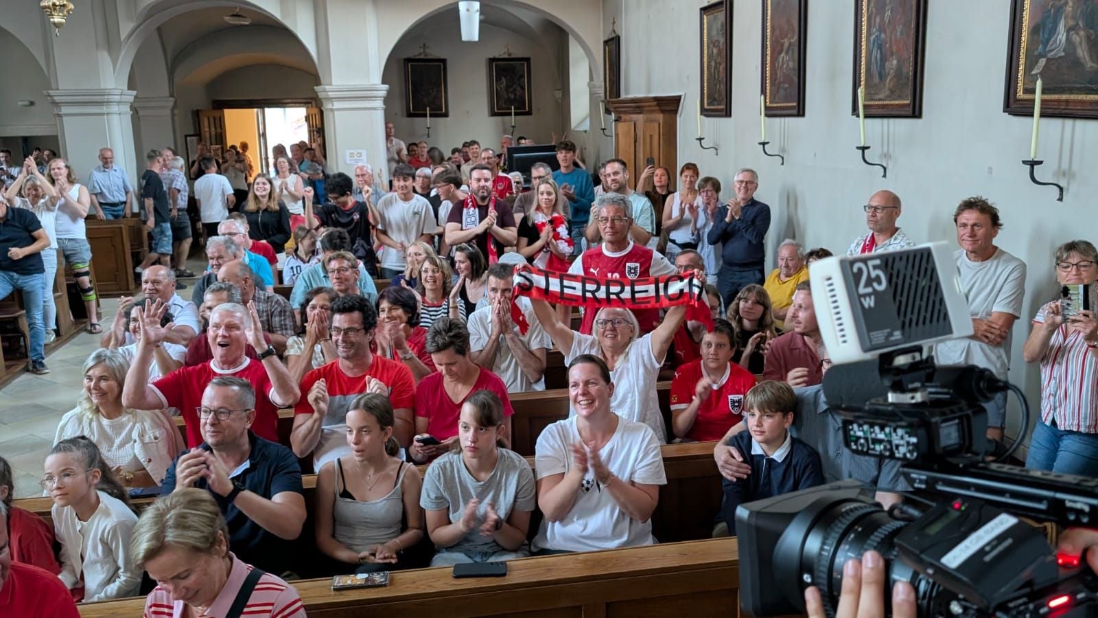 Viele Menschen jubeln und applaudieren in einer Kirche, einige tragen Österreich-Fanartikel.