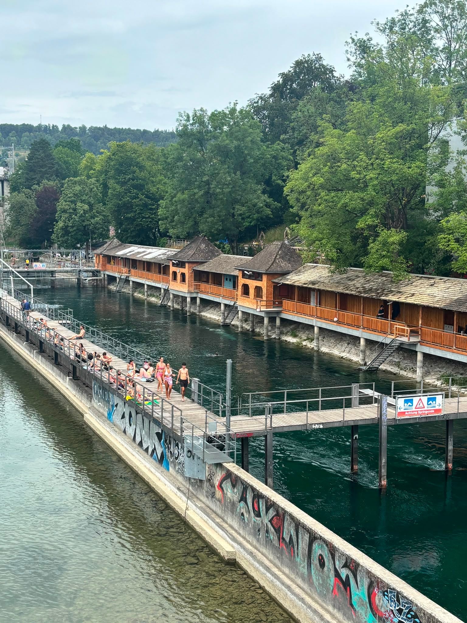 Menschen baden und entspannen am Flussbad Obere Letten bei grünen Bäumen und Holzhäusern.