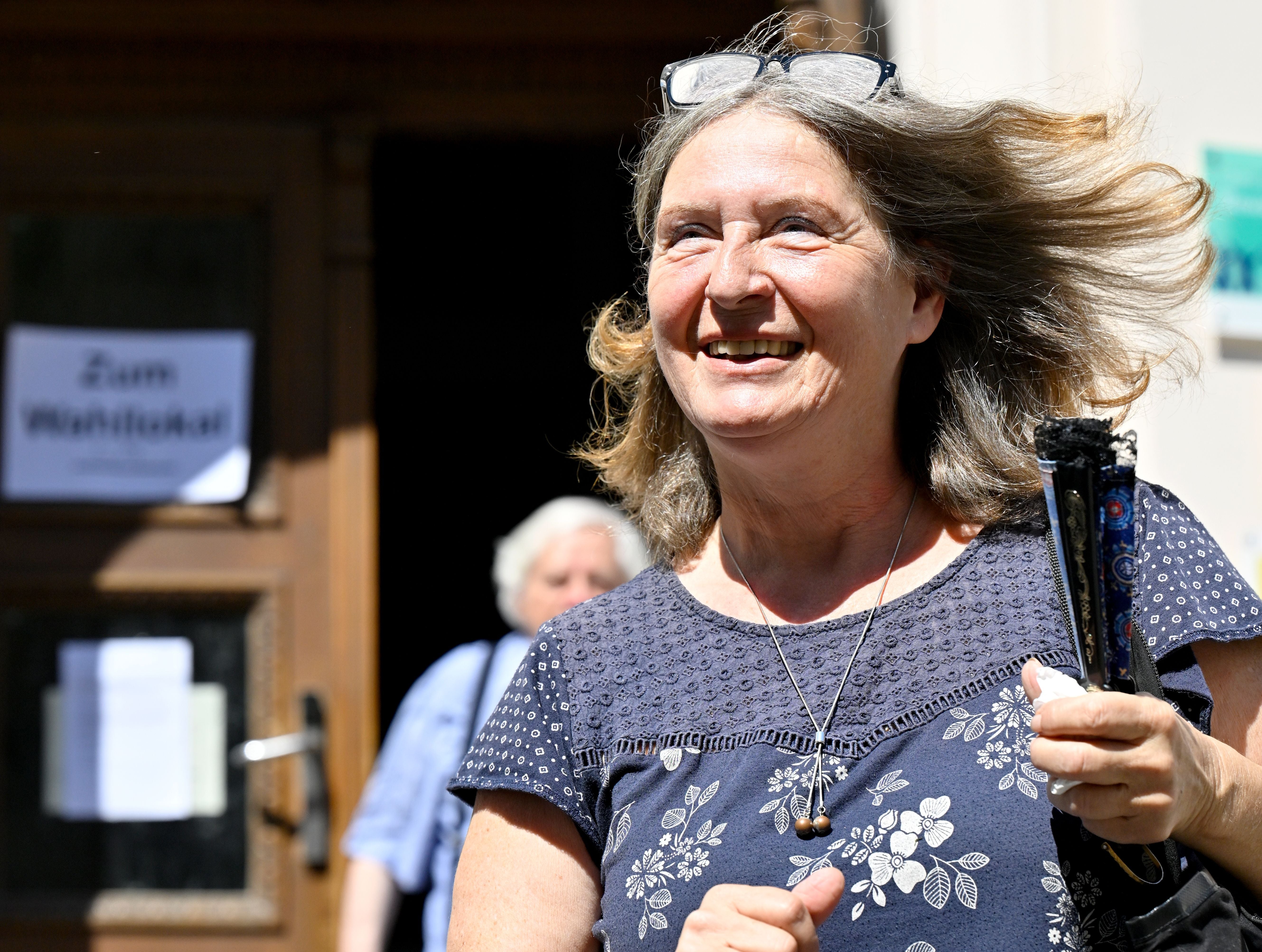 Eine Frau mit Brille im Haar lächelt vor einem Gebäude mit Wahllokalschild im Hintergrund.