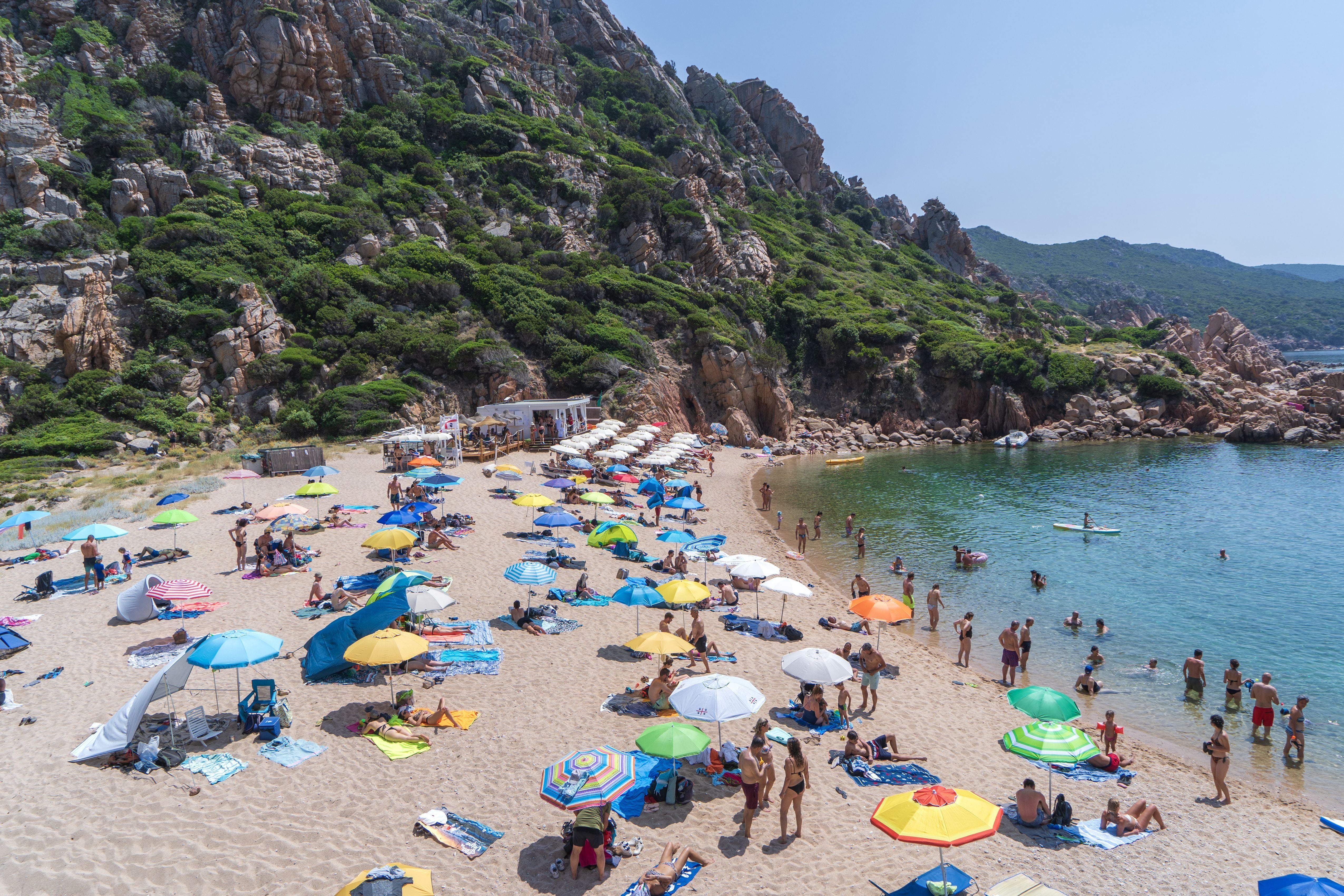 Gut besuchter Sandstrand mit bunten Sonnenschirmen, Badegästen und grün bewachsenen Felsen dahinter.