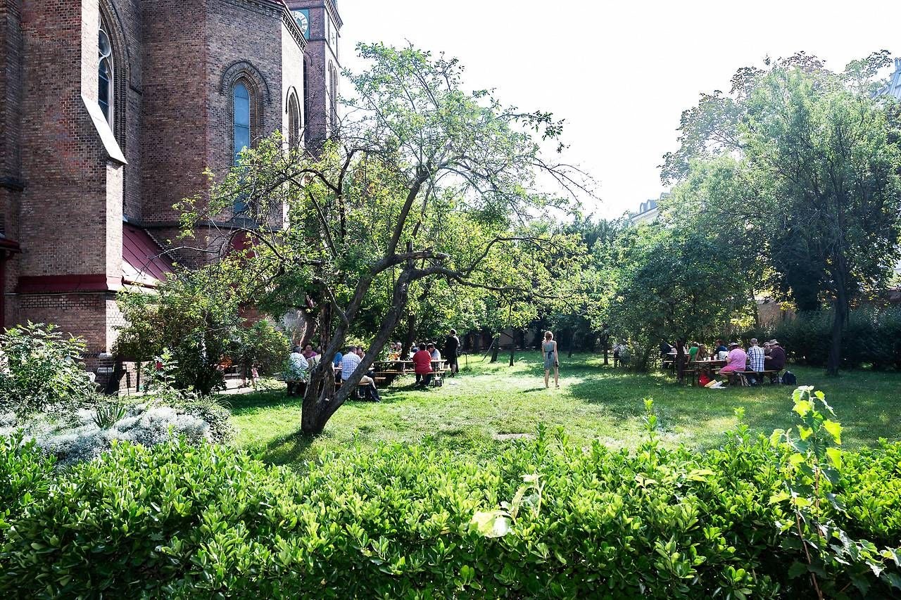 Menschen sitzen an Tischen im grünen Garten der Canisius-Pfarre am Alsergrund bei Sonnenschein.