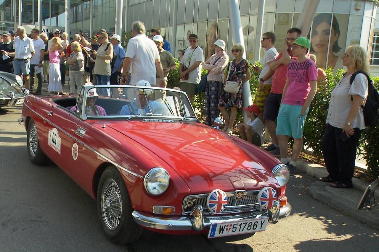 Rotes Oldtimer-Cabrio mit Union-Jack-Emblemen, umgeben von einer Menschenmenge bei Sonnenschein.