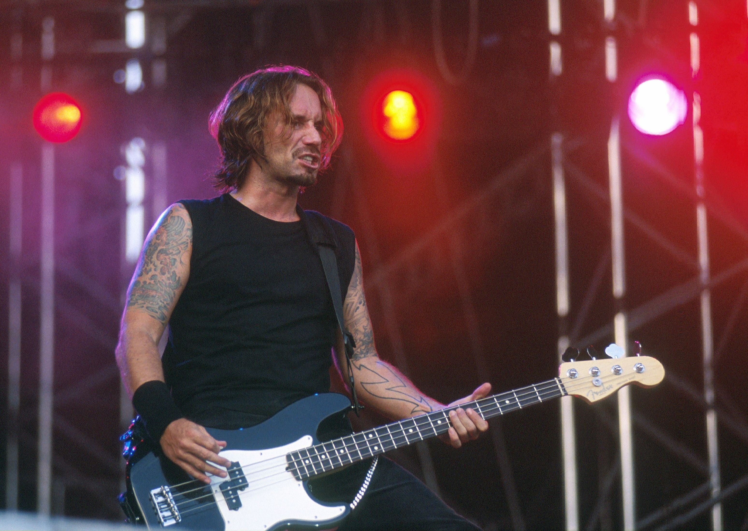 Ein tätowierter Bassist spielt bei einem Rockkonzert auf einer Bühne mit bunten Lichtern.