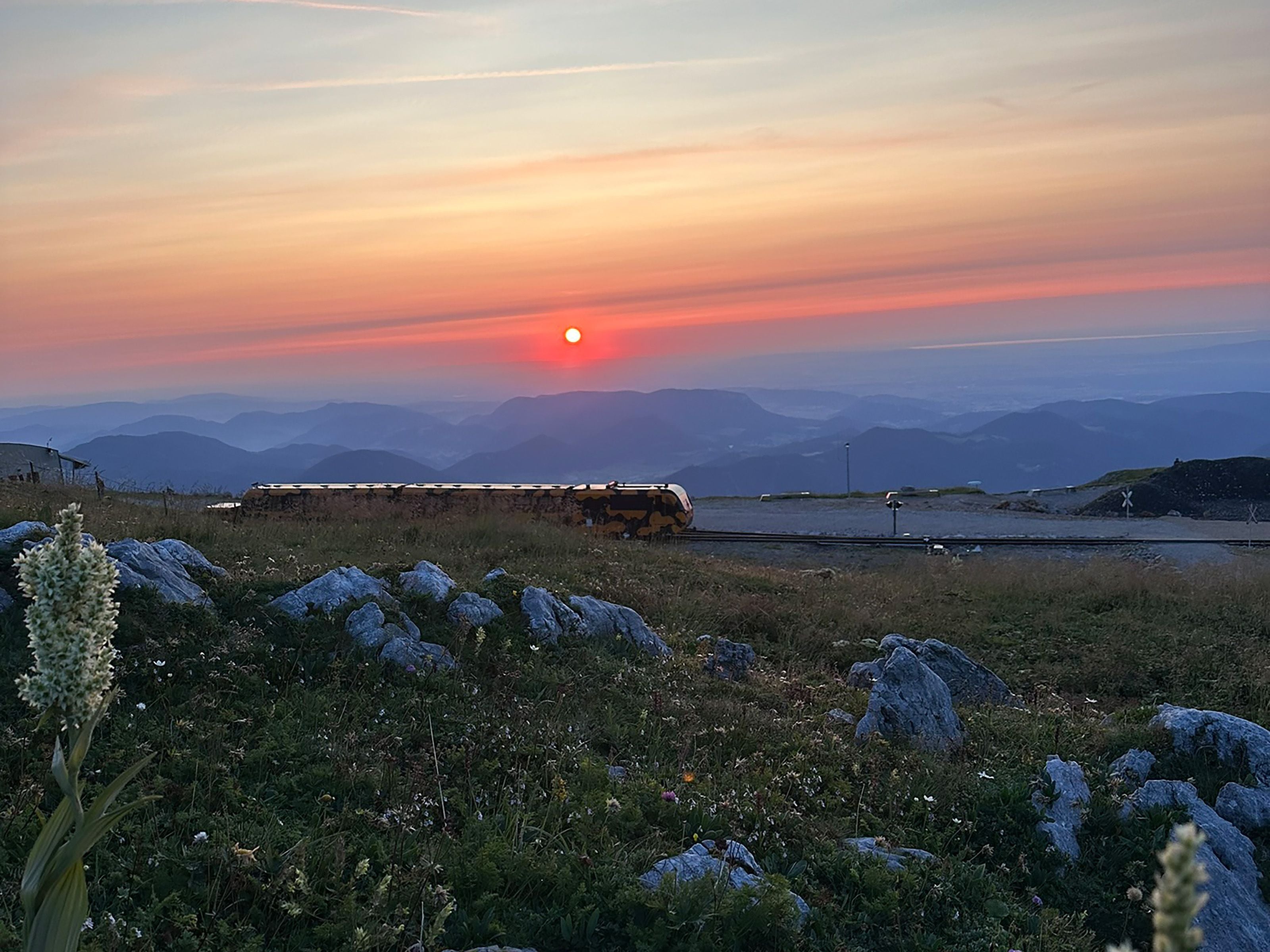 Sonnenuntergang am Schneeberg mit einer Bahn und Blick auf Berge und Wiesen im Vordergrund.
