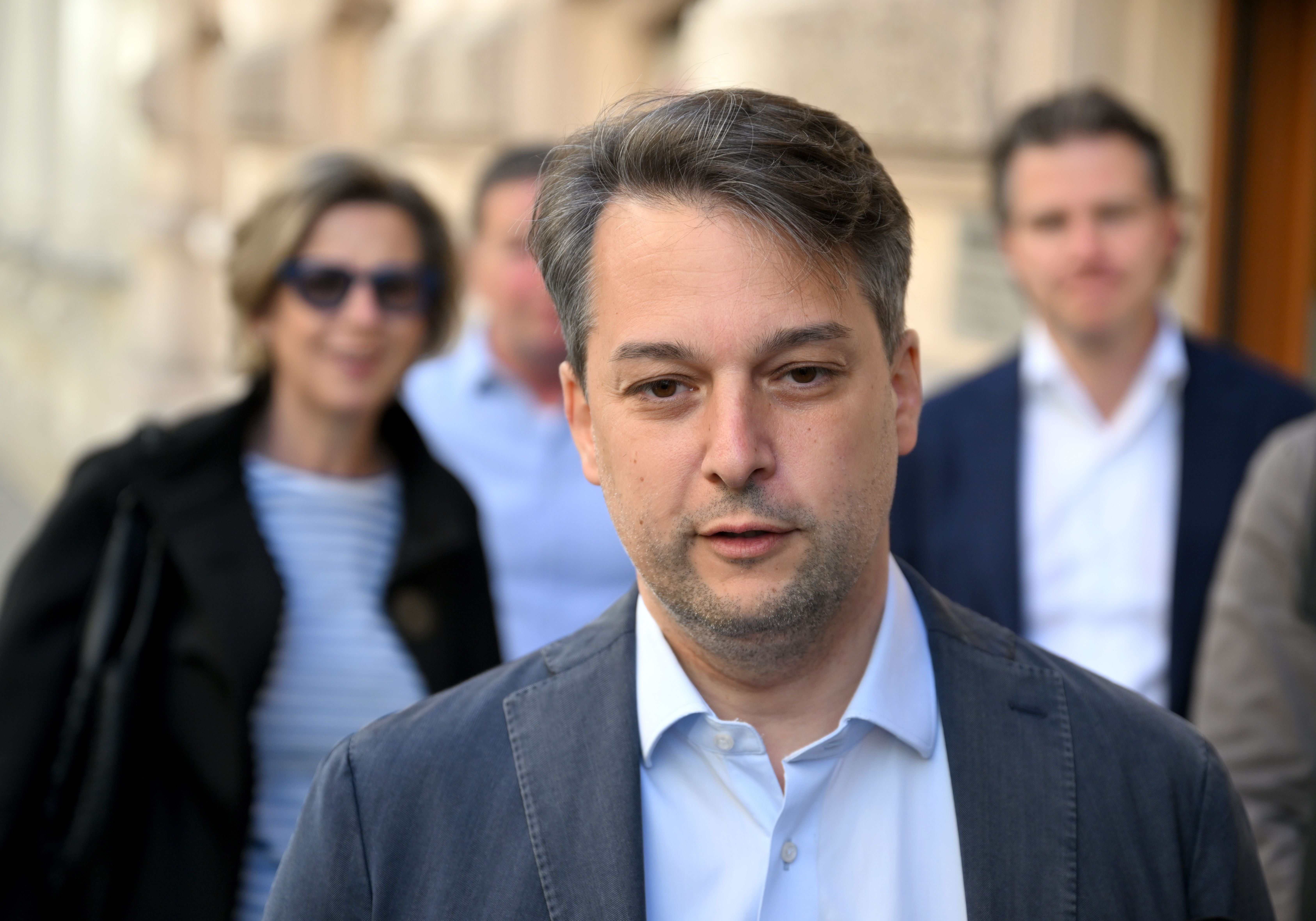 Ein Mann im Anzug steht im Vordergrund, dahinter drei unscharfe Personen draußen.