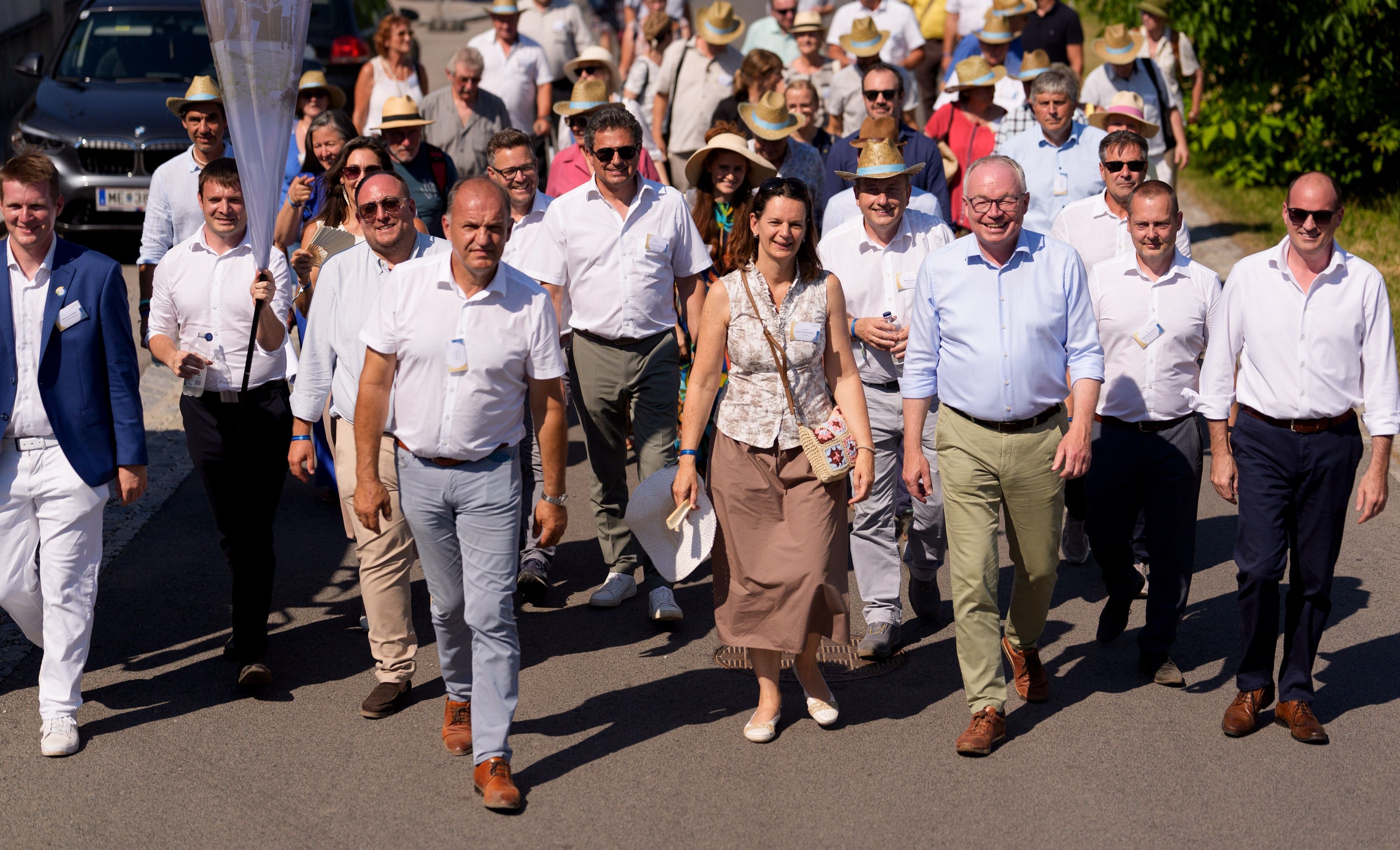 Eine große Gruppe Menschen spaziert fröhlich auf einer sonnigen Straße durch ein Dorf.