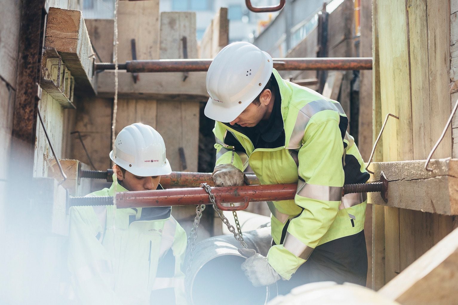 Zwei Bauarbeiter mit Helmen arbeiten an einer Baustelle mit Rohren und Holzverschalung.