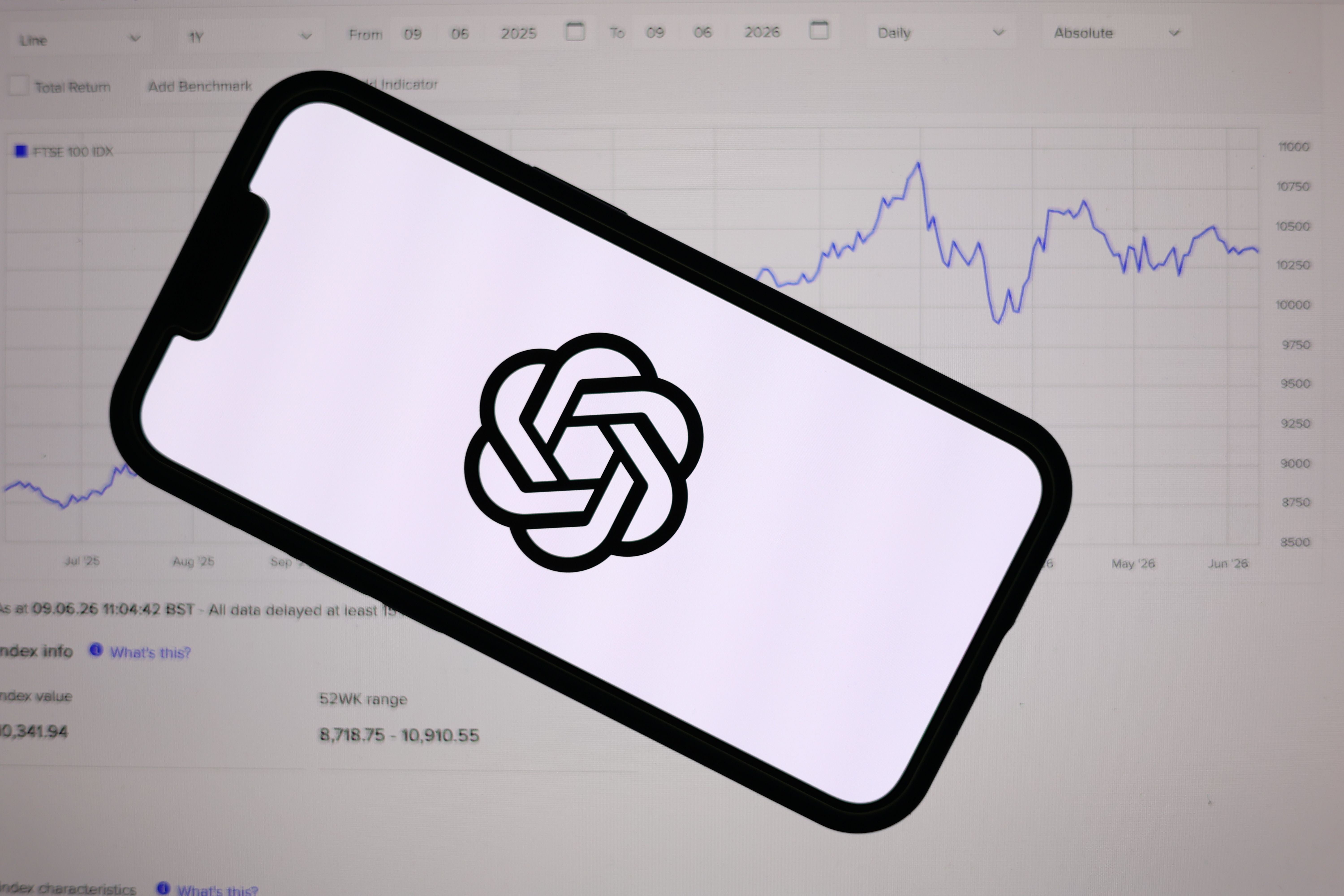 Ein Handy mit ChatGPT-Logo liegt vor einem Laptop mit Börsengrafik im Hintergrund.