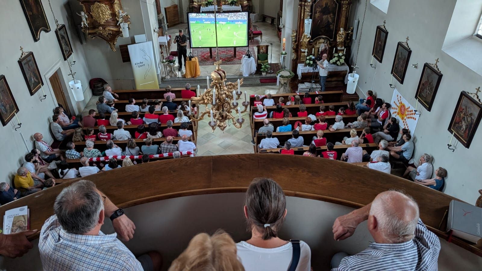 Menschen verfolgen ein Fußballspiel auf einer Großleinwand in einer Kirche von der Empore aus.