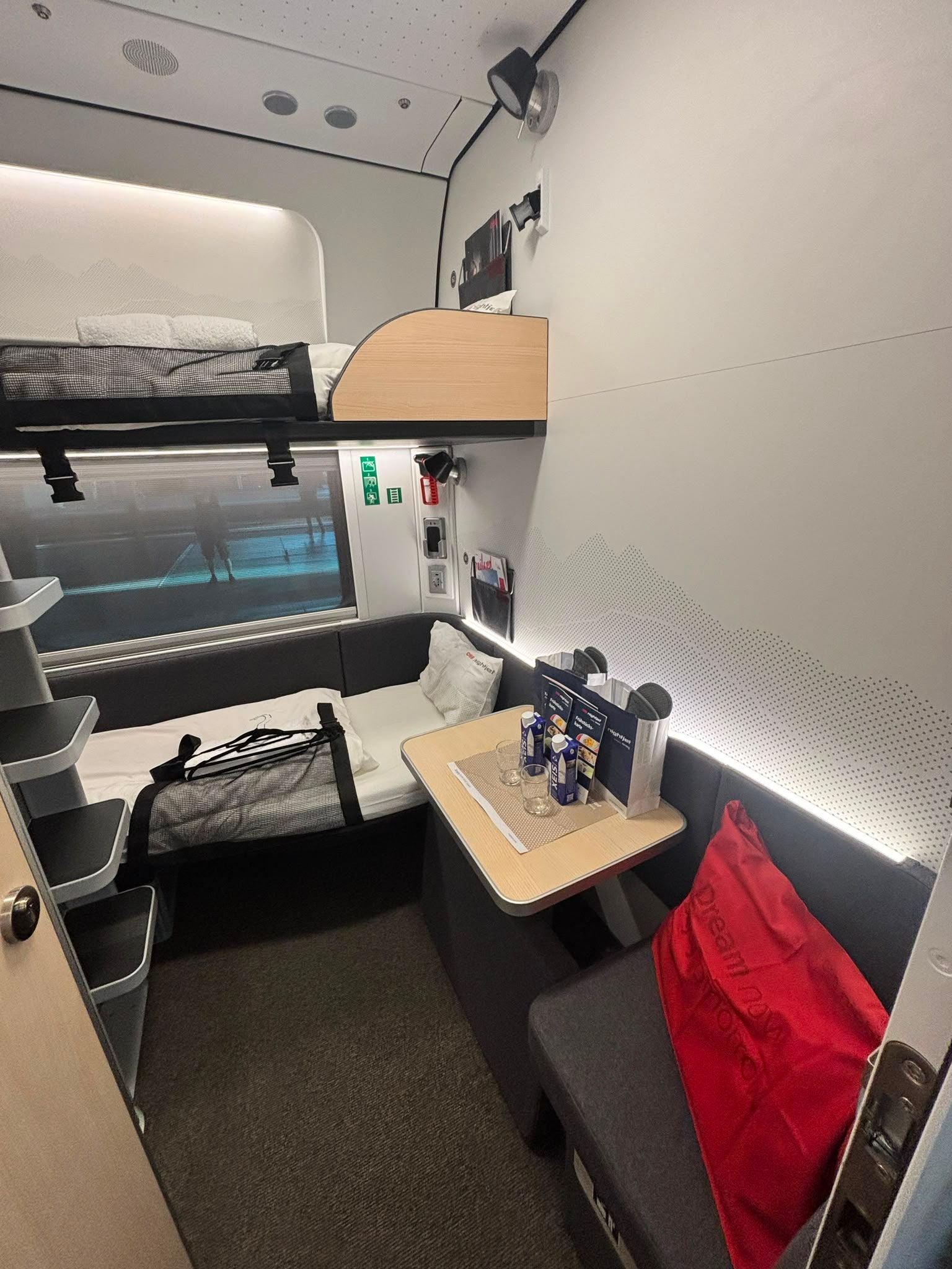 Modernes Nightjet-Schlafwagenabteil mit zwei Betten, Tisch, Sitzbank und Getränken auf dem Tisch.