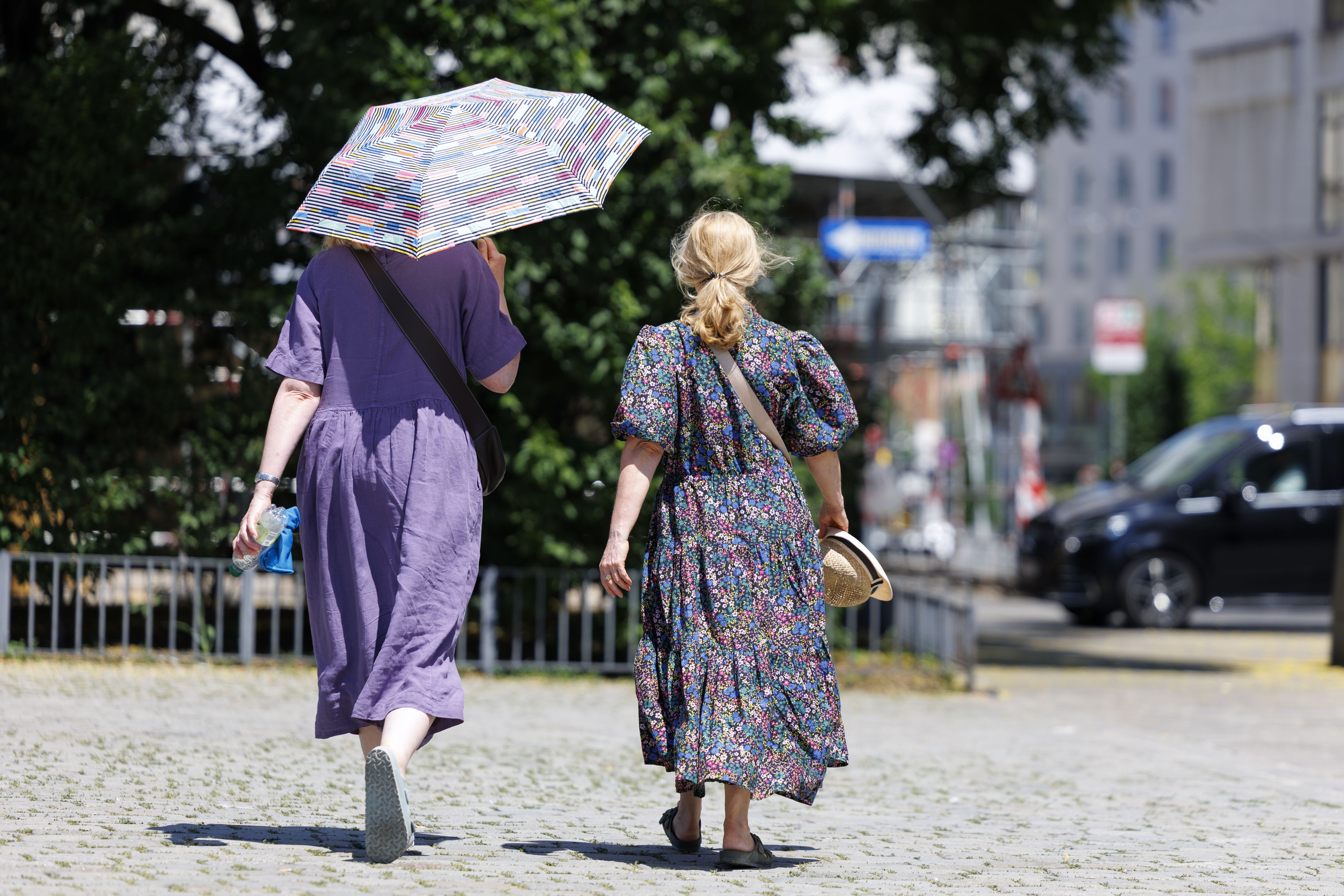 Zwei Frauen gehen im Sommer bei Hitze mit Sonnenschirm und Hut durch eine Stadt.