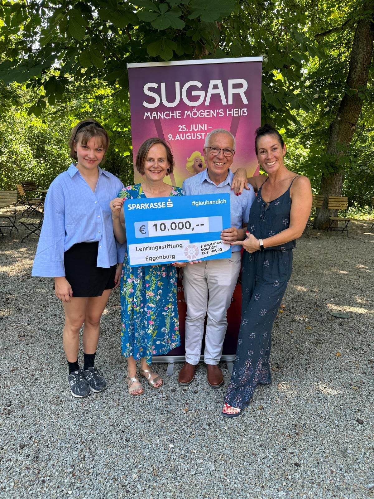 Vier Personen halten einen 10.000-Euro-Spendenscheck vor einem "SUGAR"-Plakat im Freien.