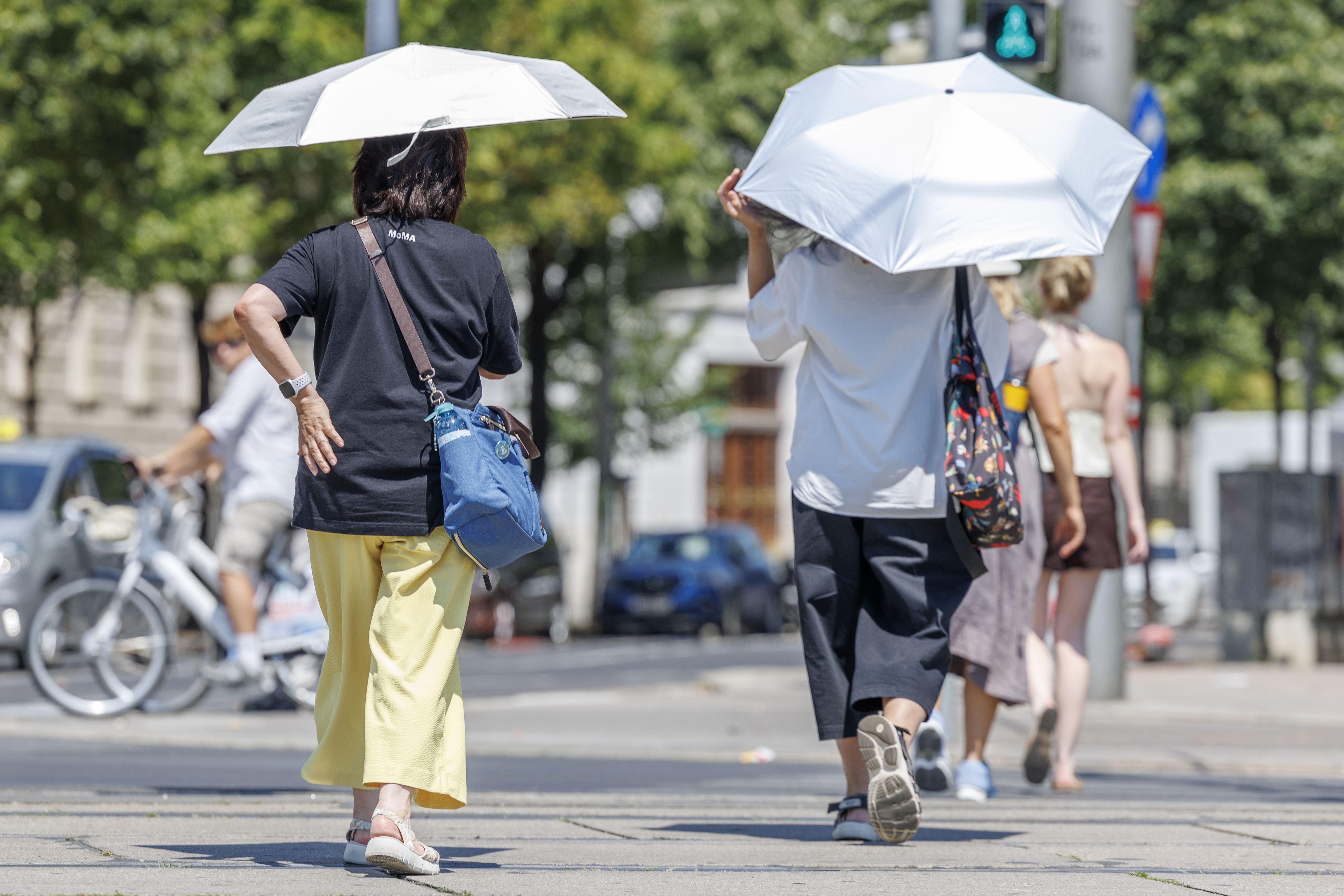 Zwei Personen gehen mit weißen Sonnenschirmen bei Sommerhitze durch eine Stadt.