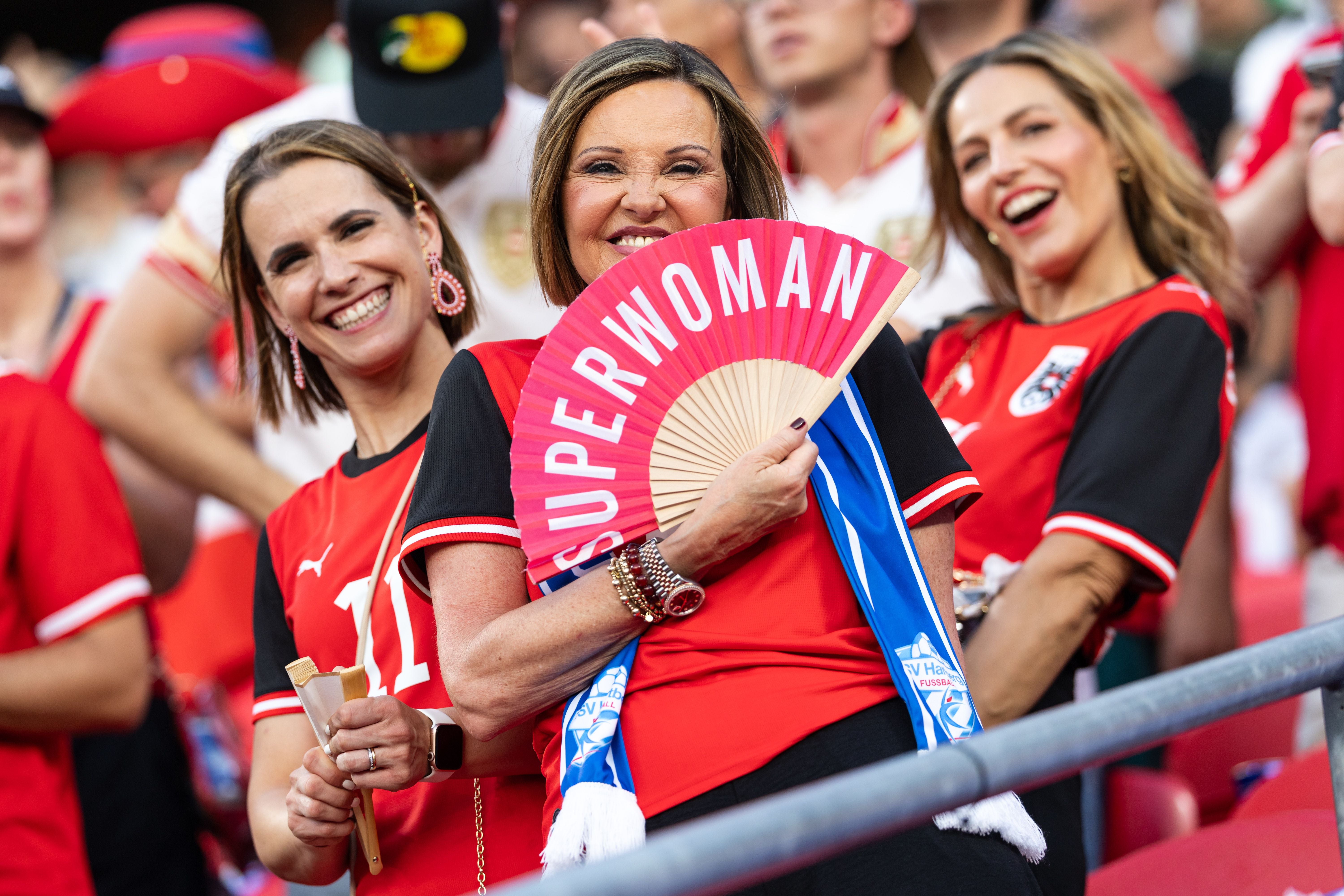 Drei fröhliche Österreich-Fans, eine hält einen Fächer mit der Aufschrift „SUPERWOMAN“.