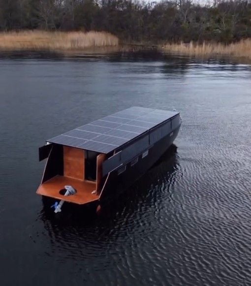 Boot mit Solarpanelen auf dem Dach fährt auf einem ruhigen See, im Hintergrund Schilf und Bäume.
