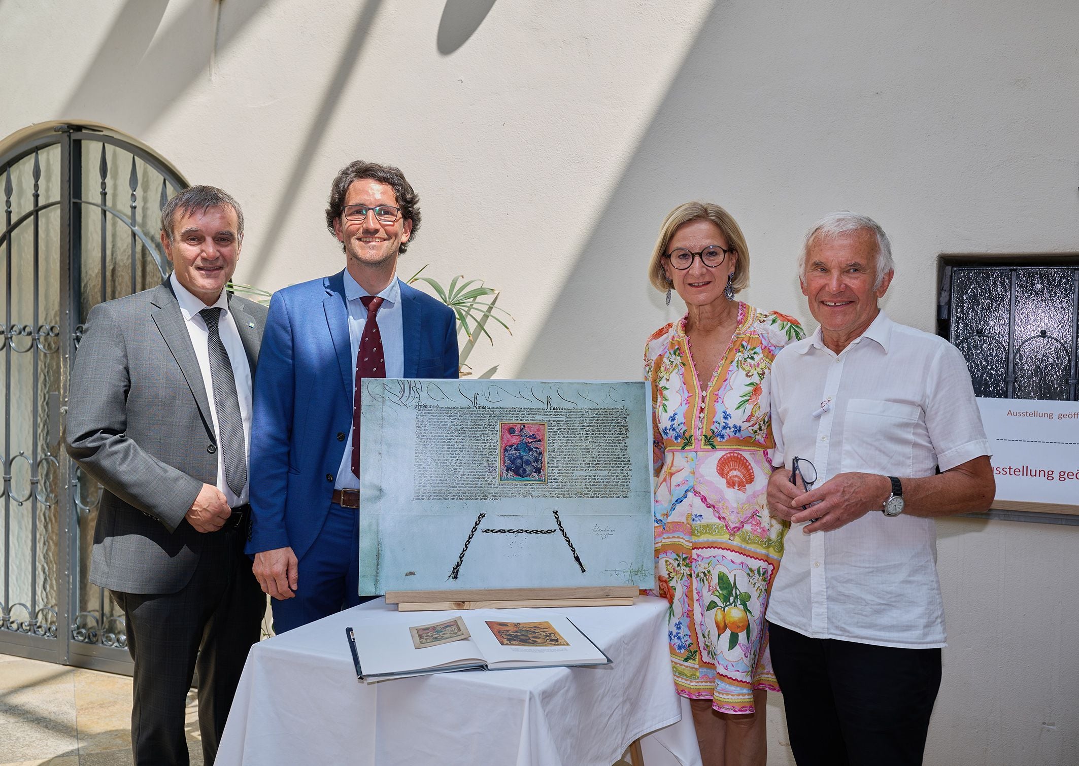 Vier Personen stehen bei einer Ausstellung neben einer großen Urkunde auf einem Tisch.