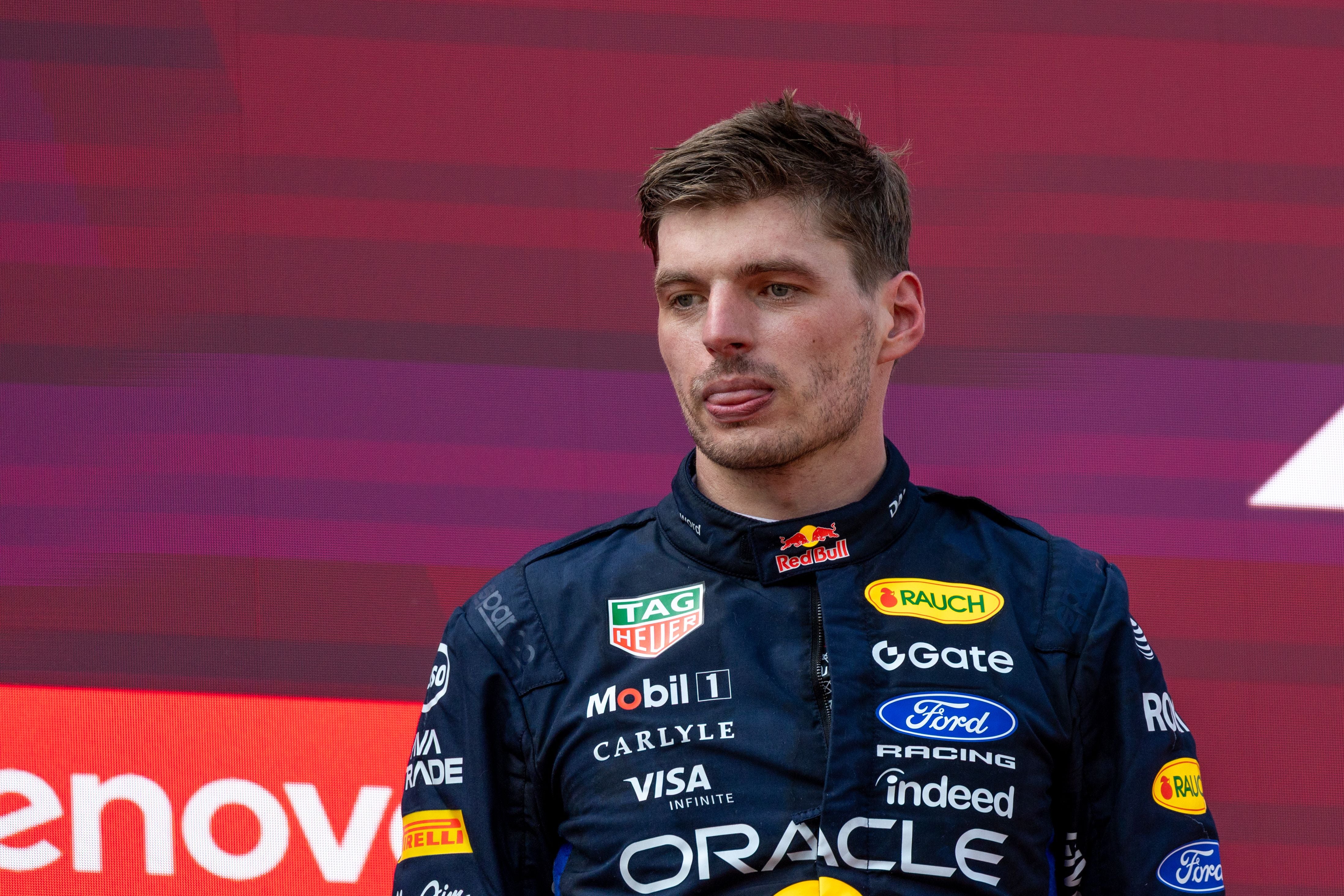 Ein Rennfahrer im Red-Bull-Anzug steht vor einer roten Kulisse und streckt die Zunge heraus.