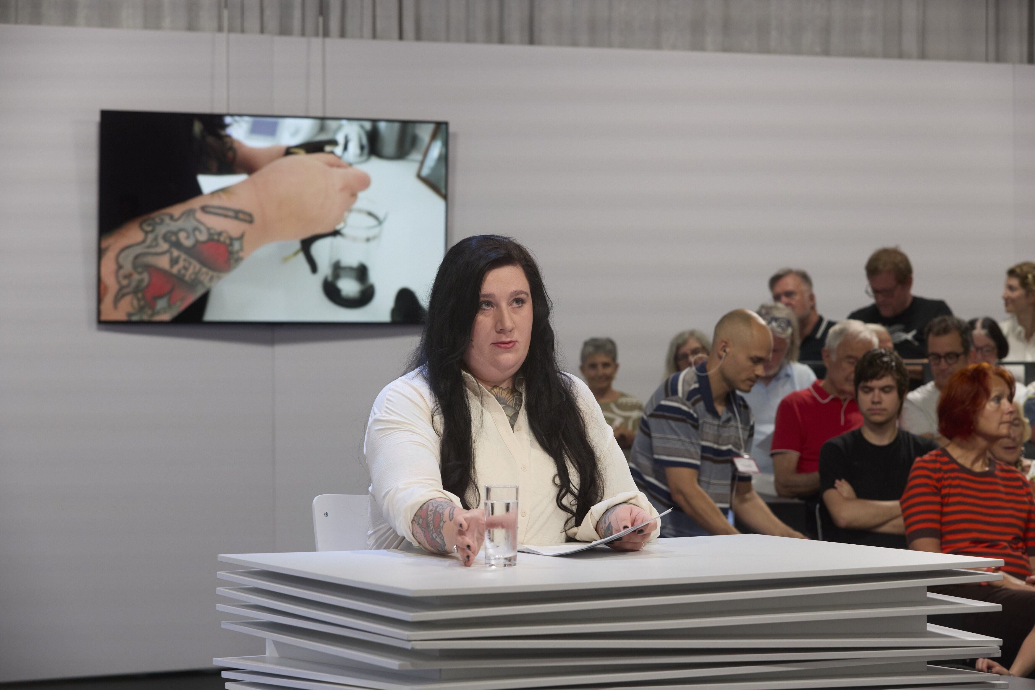 Frau mit langen dunklen Haaren liest bei einer Veranstaltung, Publikum im Hintergrund.