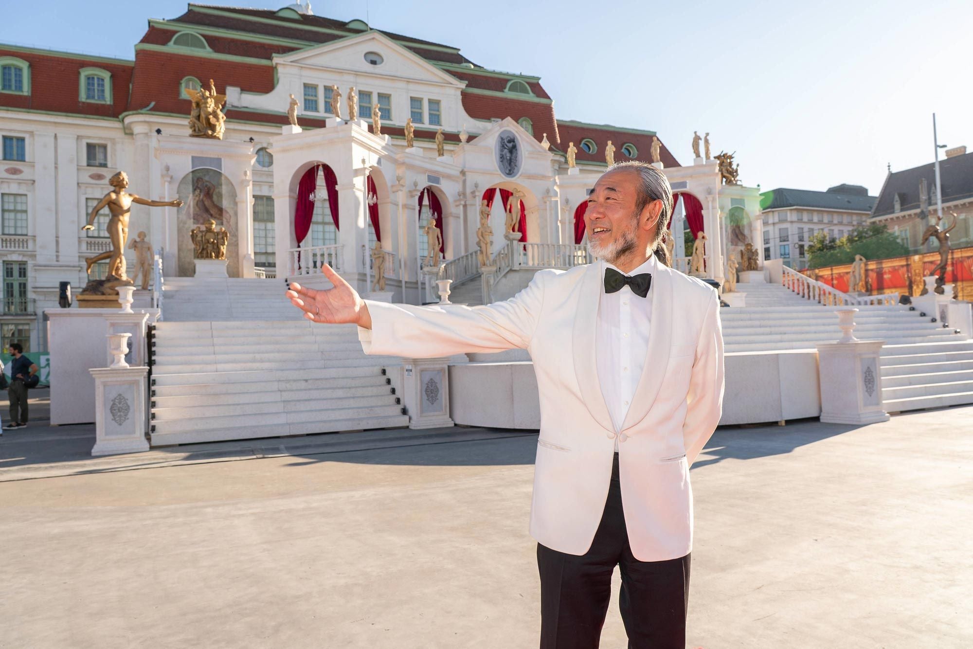 Ein elegant gekleideter Mann steht vor einem prunkvollen Opernhaus mit roten Vorhängen.